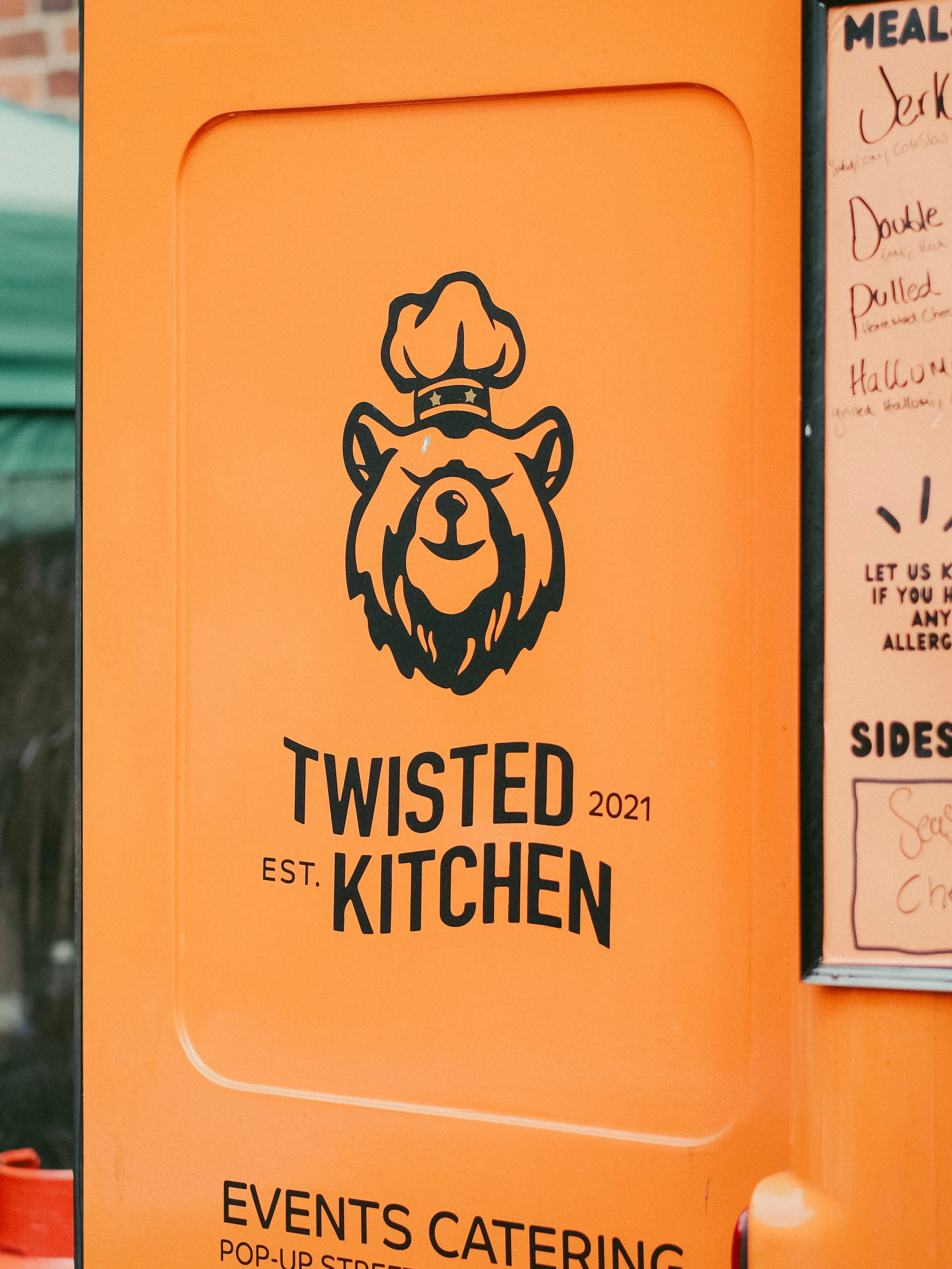 Orange sign with a logo of a bear wearing a chef's hat and the text 'Twisted Kitchen 2021 Est. Twisted Kitchen'.