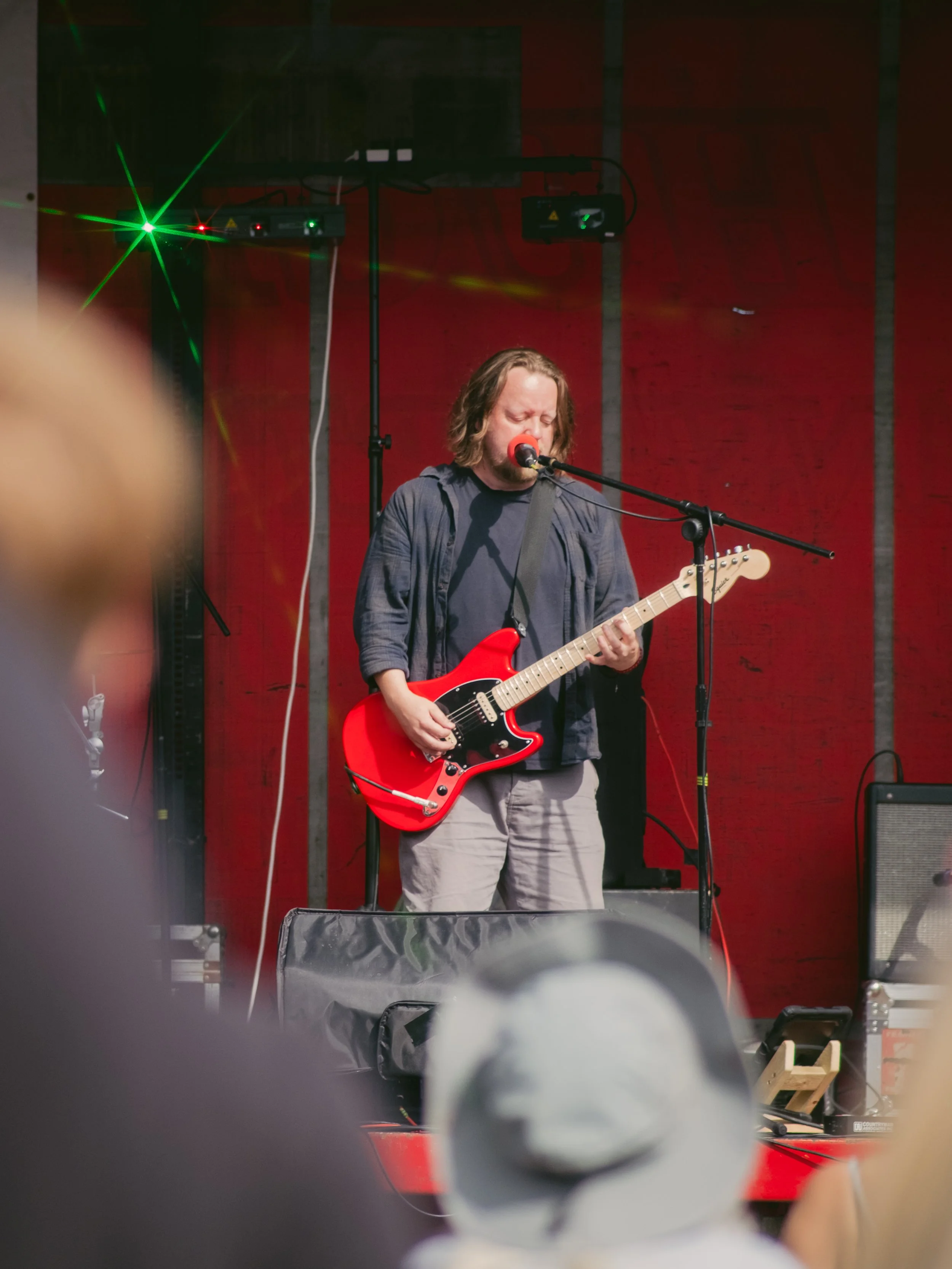 Man with shoulder-length hair singing into a microphone and playing an electric guitar on stage with a red background and green laser lights.