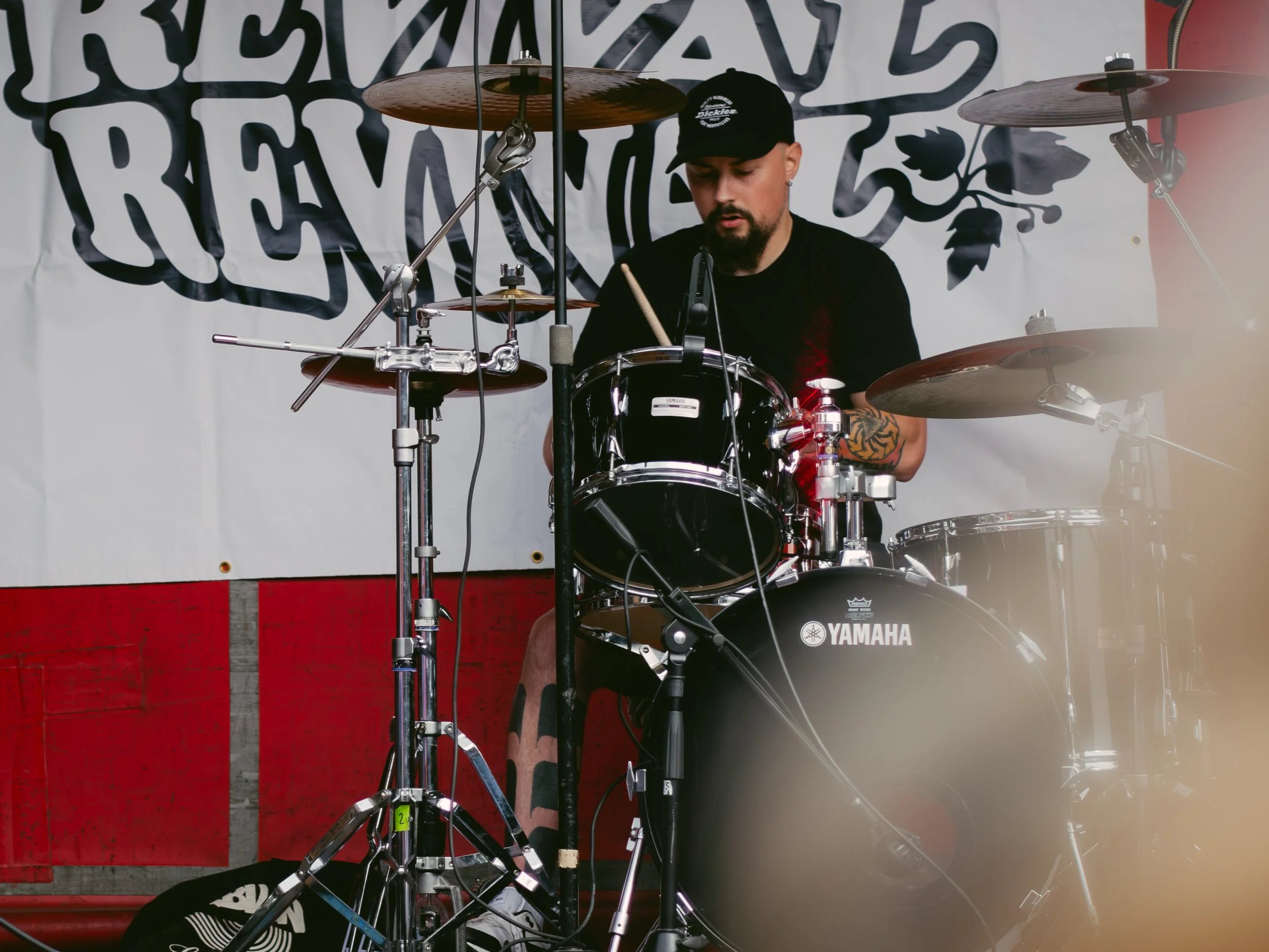 A man playing a black Yamaha drum set on stage with a white background featuring black text and a design, wearing a black baseball cap and black t-shirt.