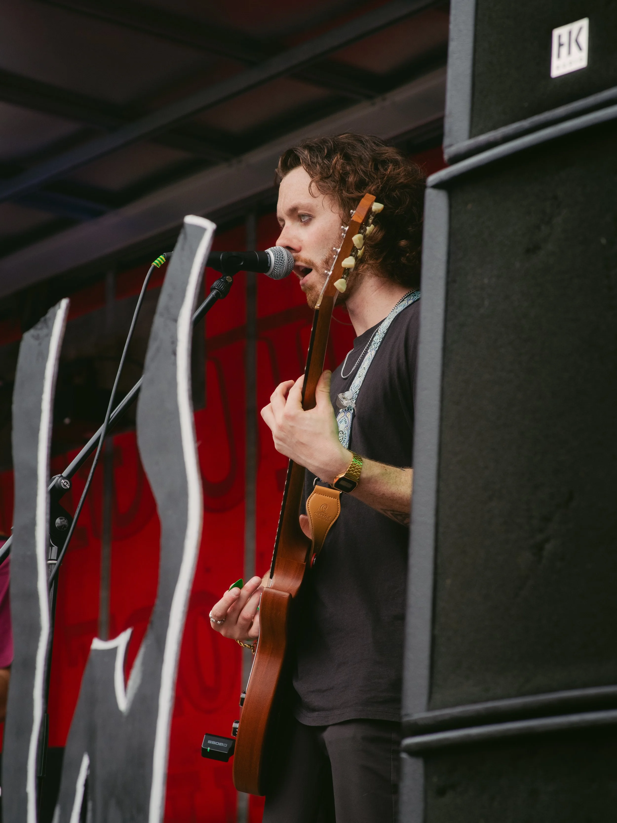 A male musician singing into a microphone while playing an acoustic guitar, on stage with a red backdrop, partially obscured by large black speakers.