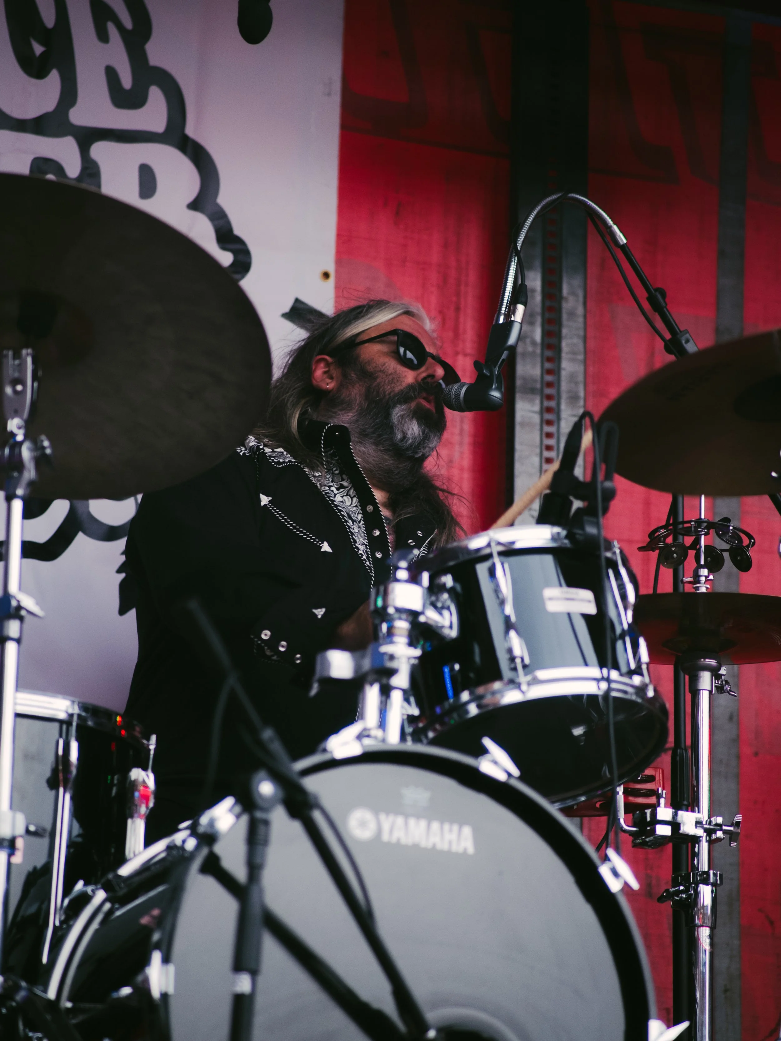 A male drummer with long hair, beard, and sunglasses playing a Yamaha drum set with a microphone in front of him during a live performance.