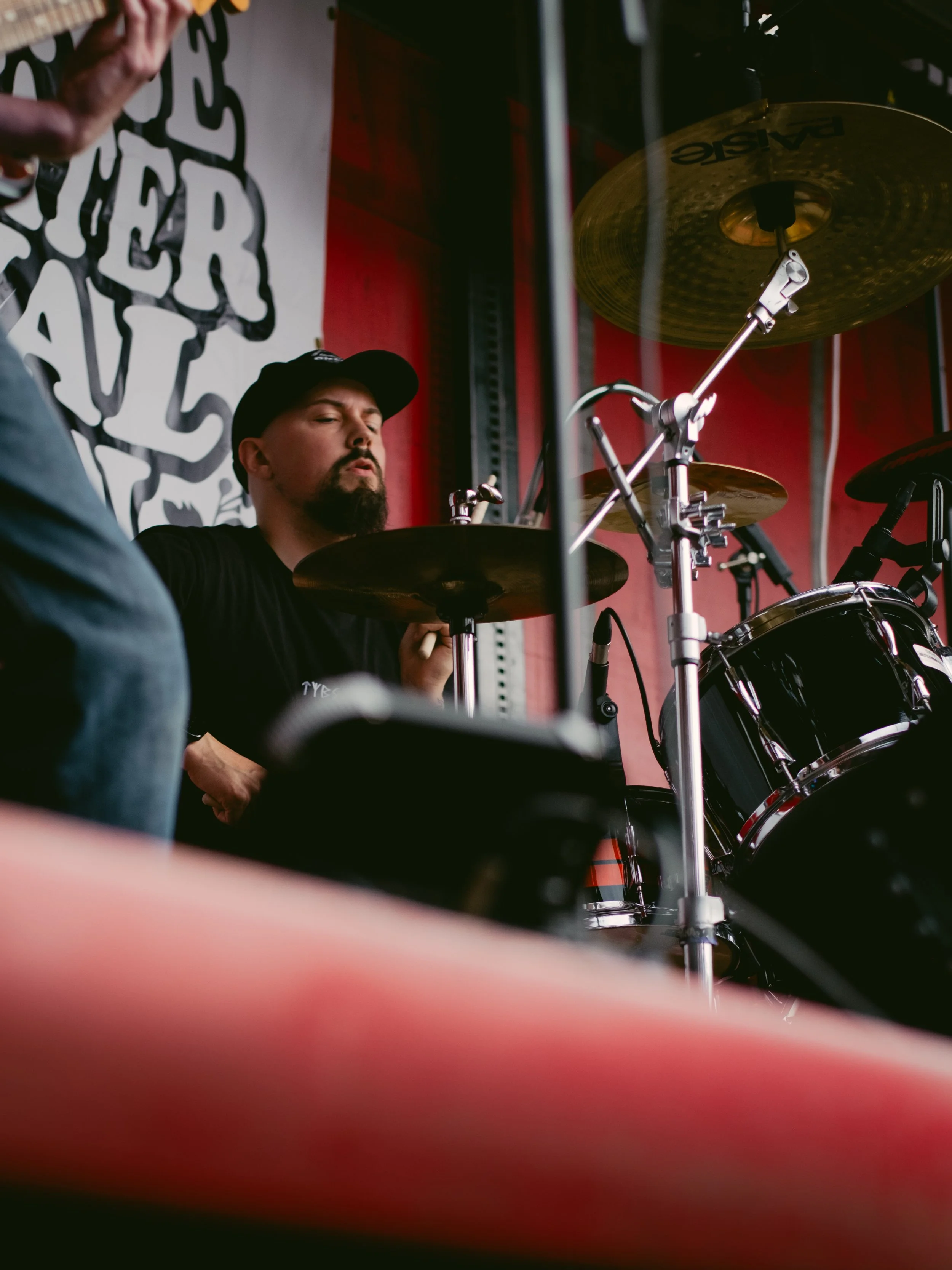 A man with a beard and black cap playing drums during a live performance, with a red background and a black and white banner behind him.