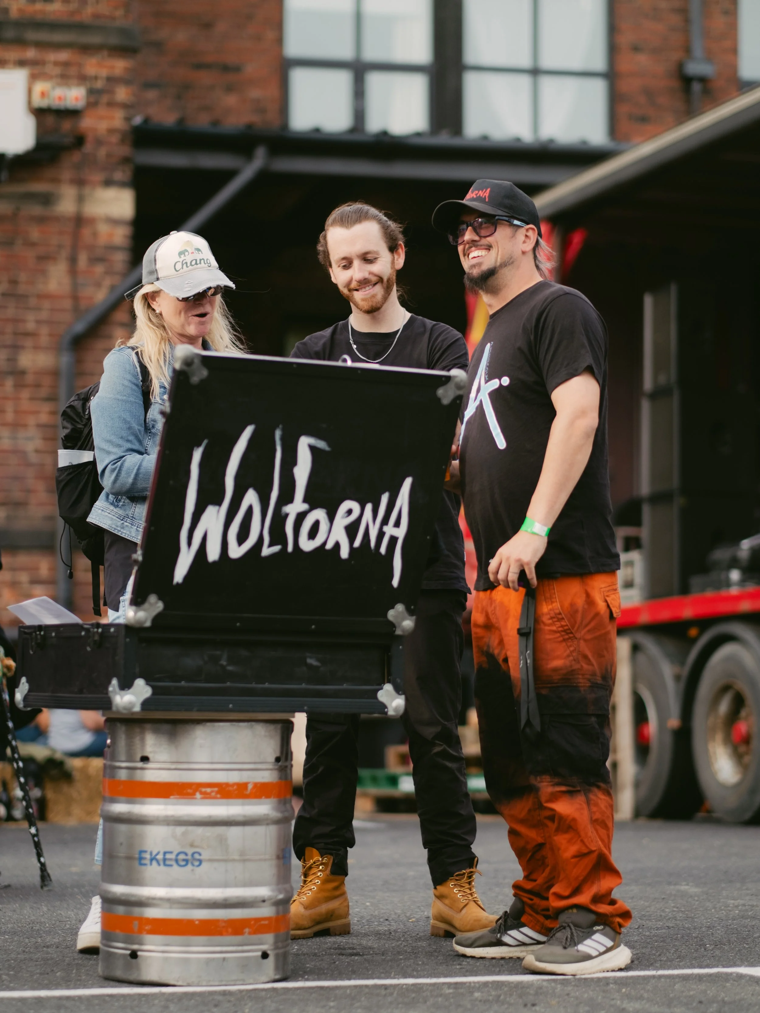 Three people smiling and looking at a black sign with the word 'Woltorna' on it, set on a metal barrel, in an outdoor setting with a brick building in the background.