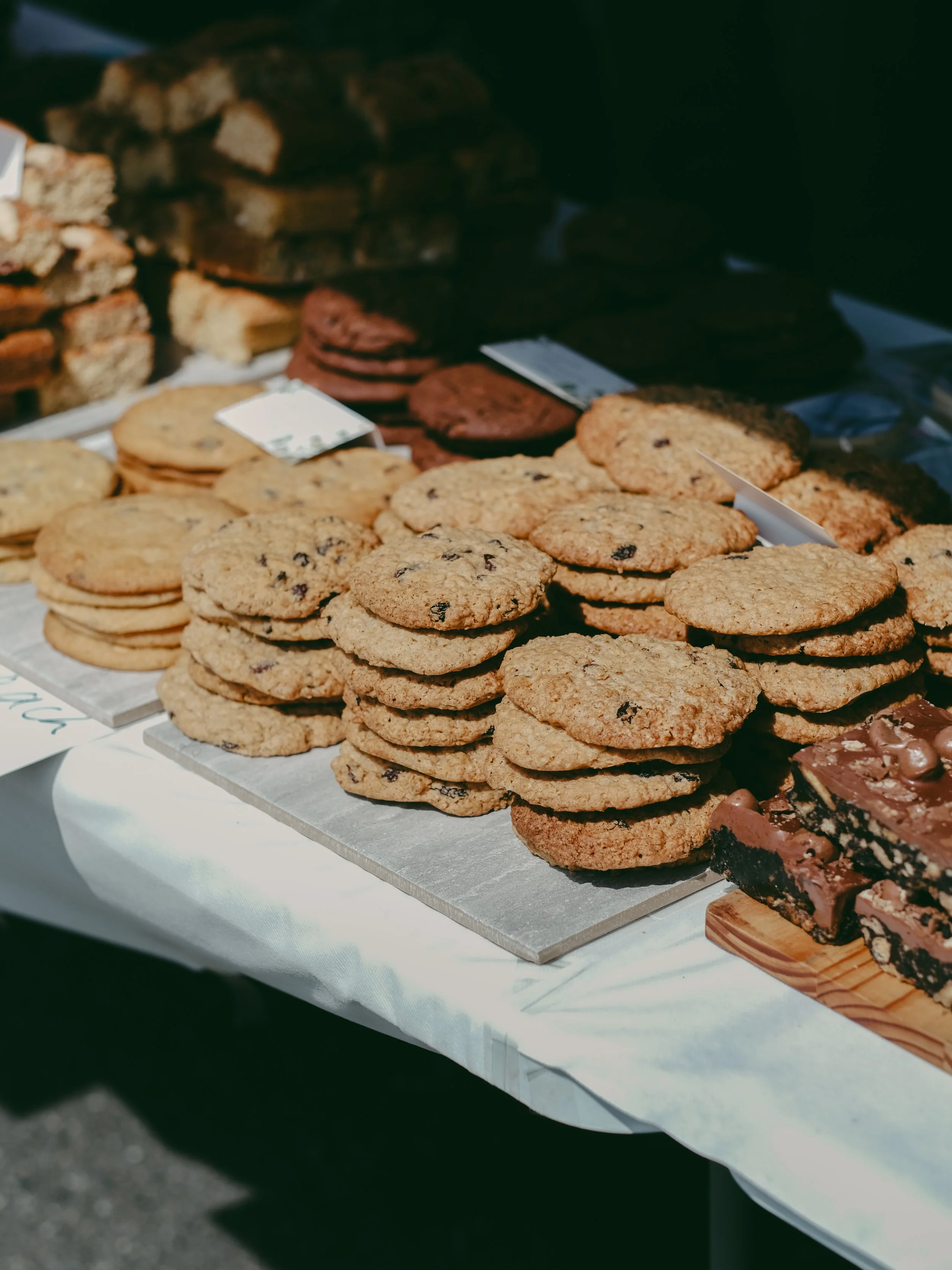 A table with stacks of chocolate chip cookies, placing on parchment paper, and some brownies on a wooden board, with other cookies and baked goods in the background.