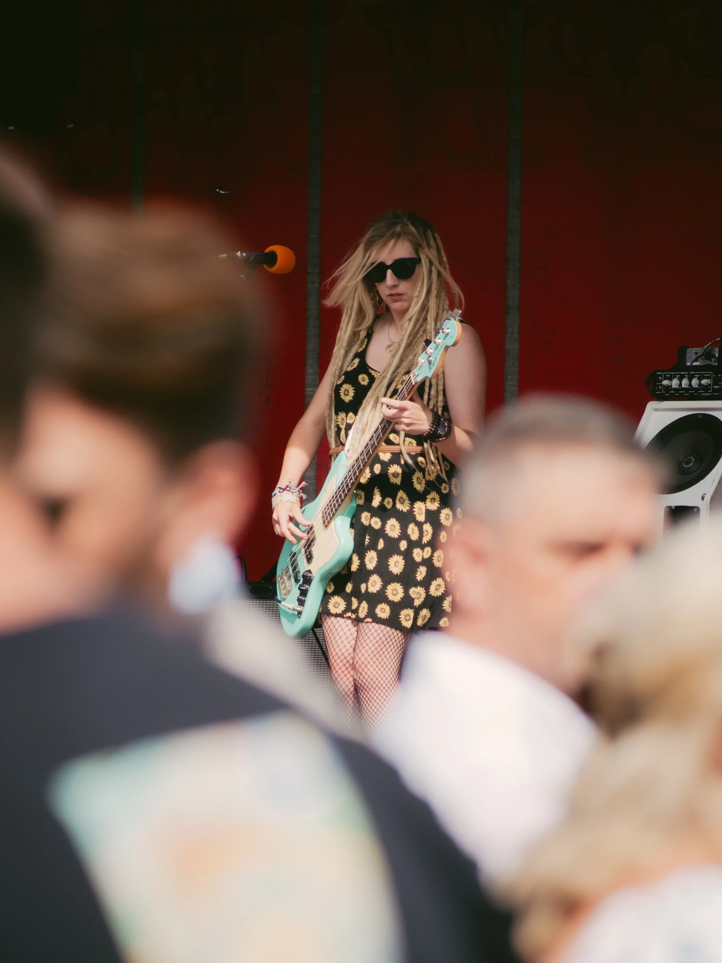 A young woman with dreadlocks, wearing sunglasses, a sunflower-patterned dress, fishnet stockings, and wristbands, playing a blue electric bass guitar on stage during a live music performance.