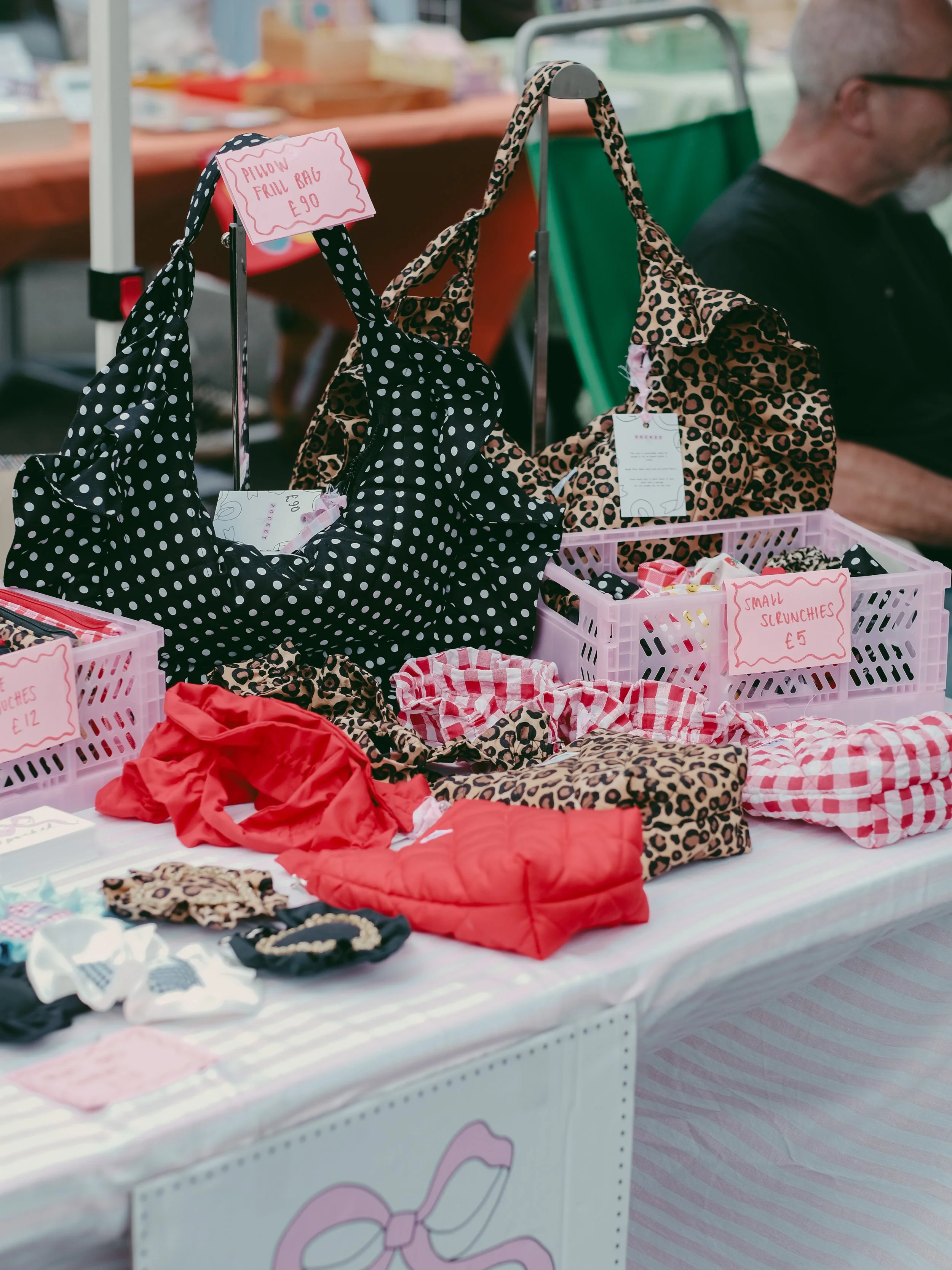 A market stall displaying polka dot and leopard print bags, assorted fabric items, and signs indicating prices for various accessories, with a man sitting behind the stall.