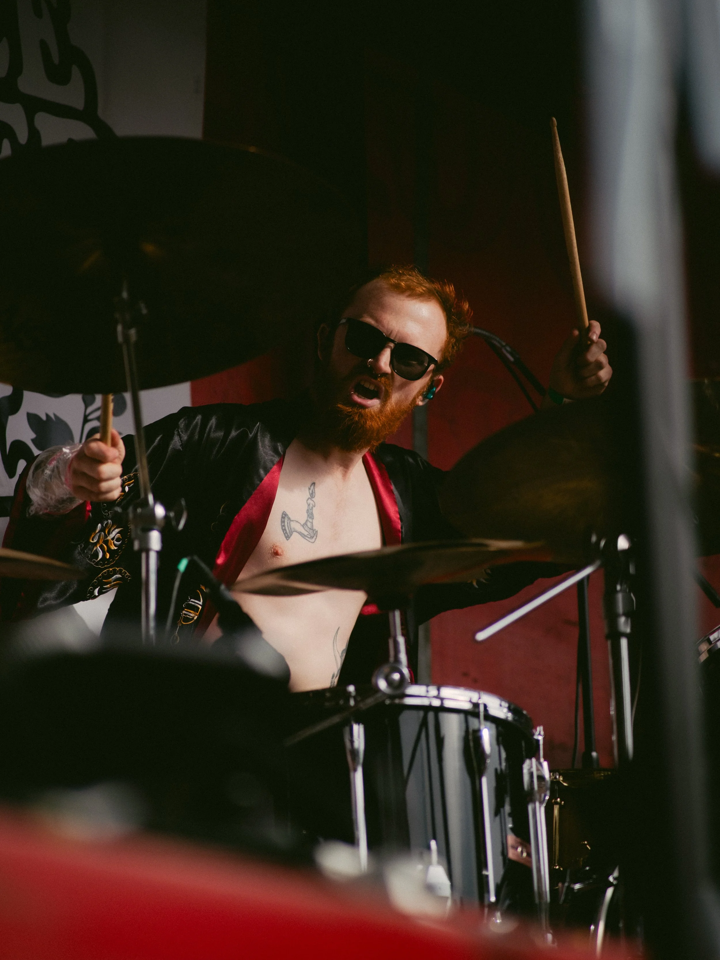 A male drummer with red hair and a beard, wearing sunglasses and a black and red jacket, playing drums passionately on stage.