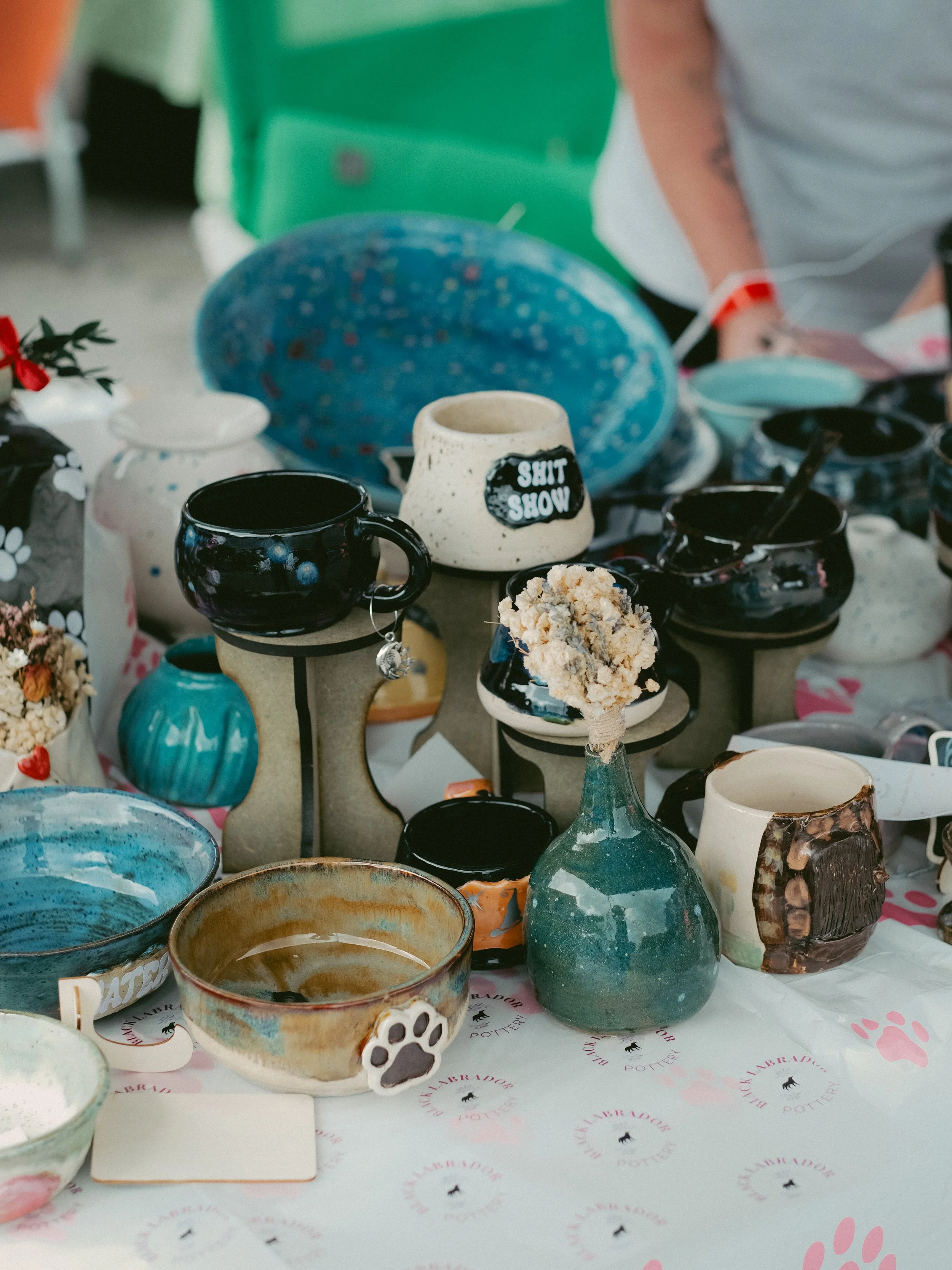 Display of various handmade pottery items, including mugs, bowls, and vases, on a table at a pottery market or show.