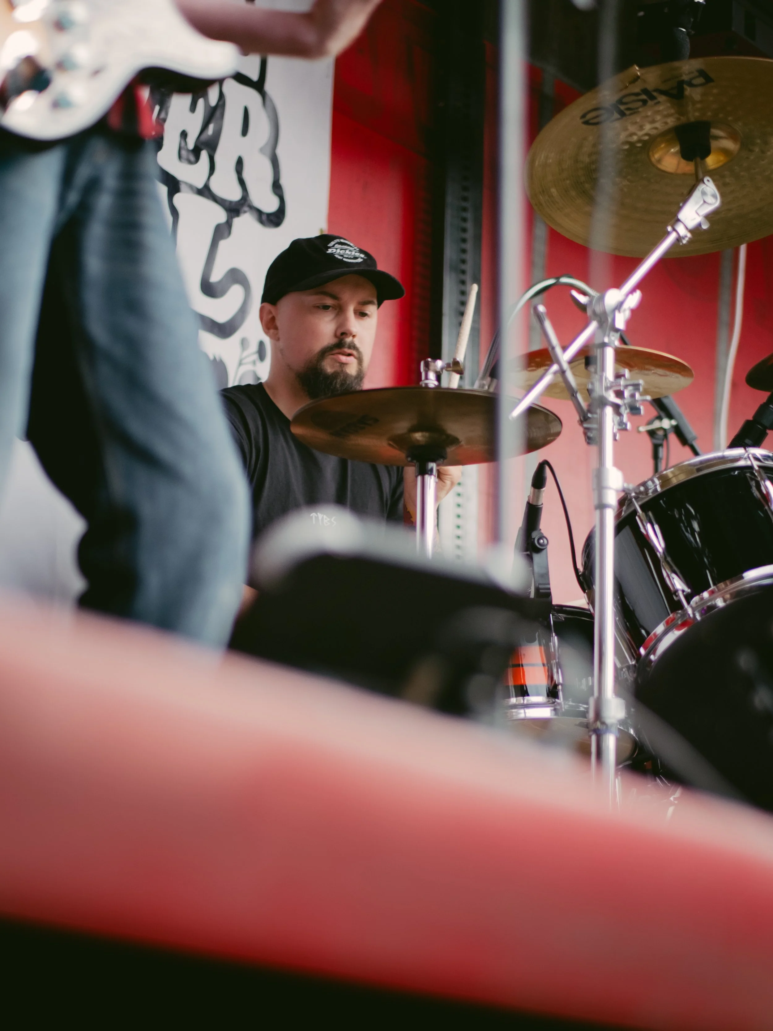 A man playing a drum set during a performance at a music venue with a red wall and a logo in the background.