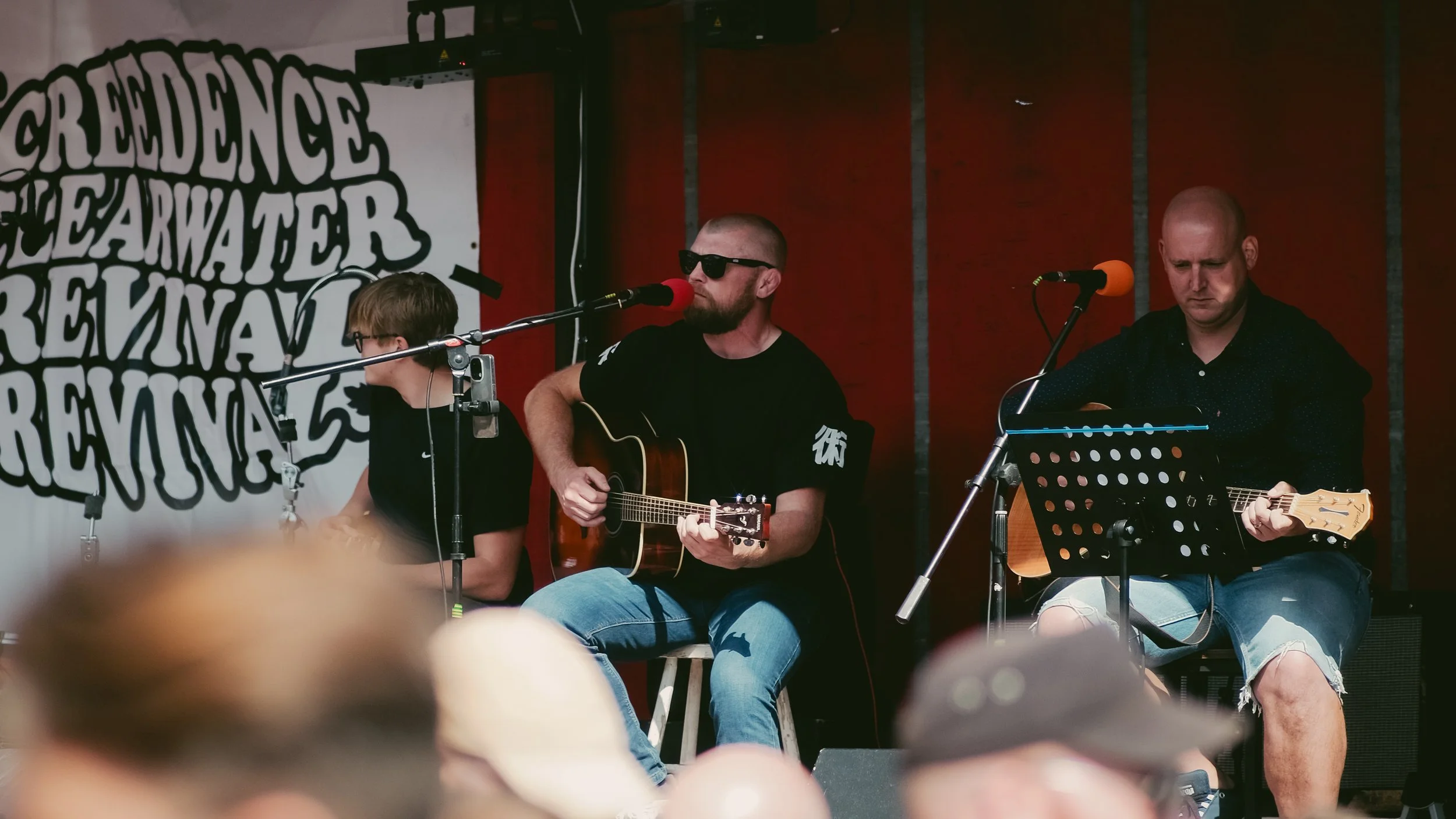 Three musicians performing on an outdoor stage; two are playing acoustic guitars, one is singing into a microphone, and music stands are visible.