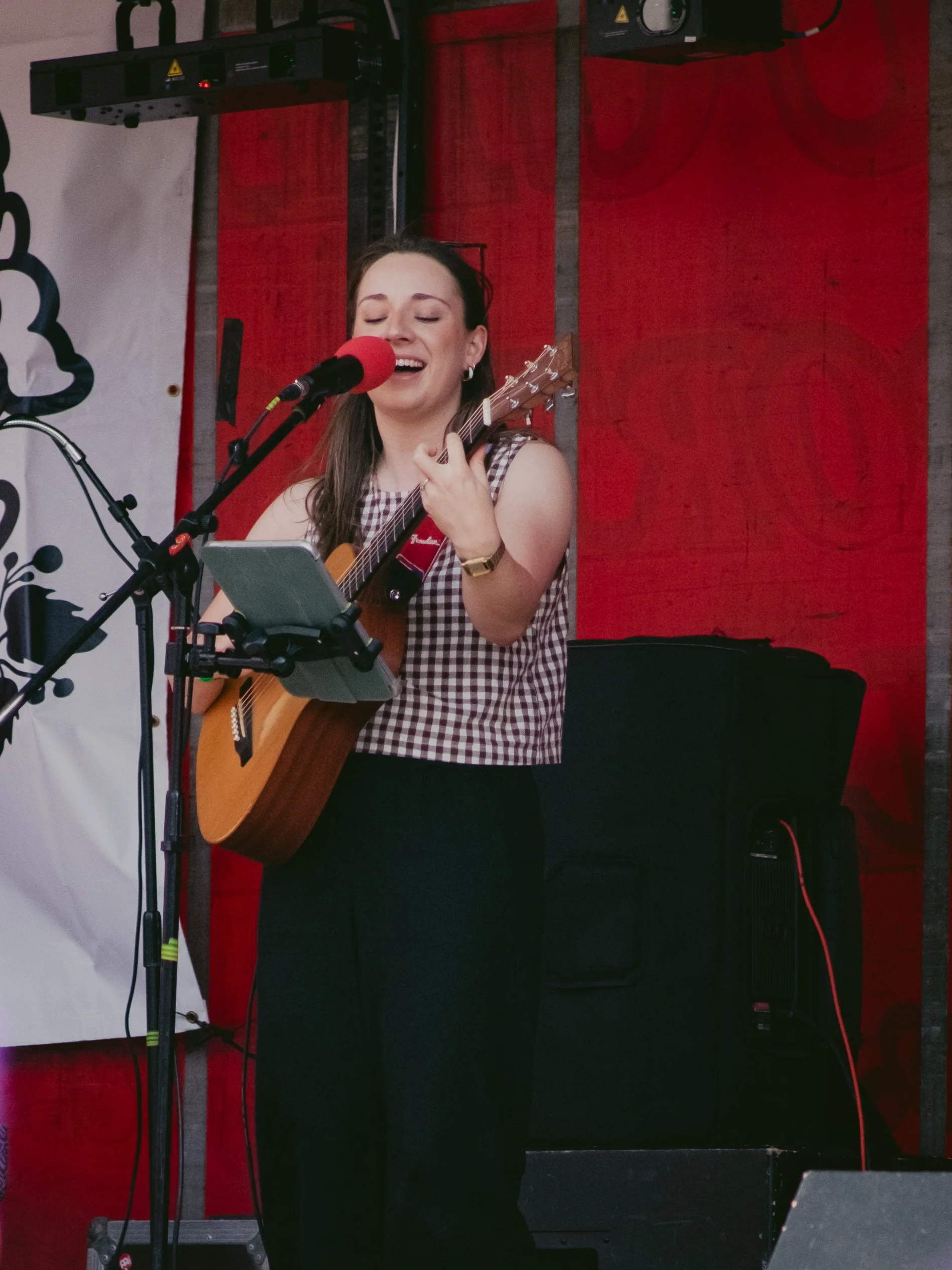 A woman performing music on stage, singing into a microphone and playing an acoustic guitar, with her eyes closed and smiling.