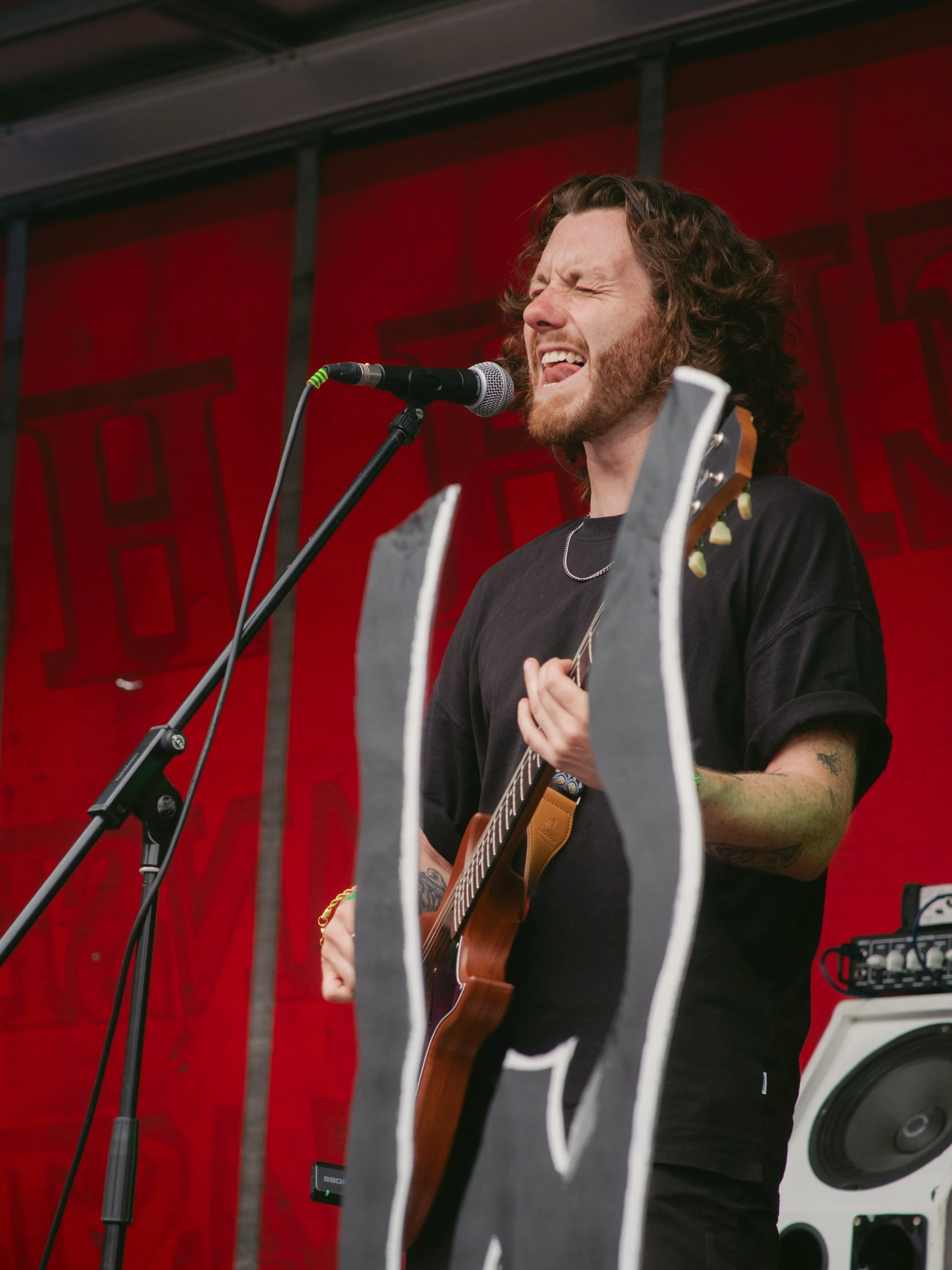 Male musician with curly hair and tattoos singing passionately into a microphone and playing an acoustic guitar during a performance.