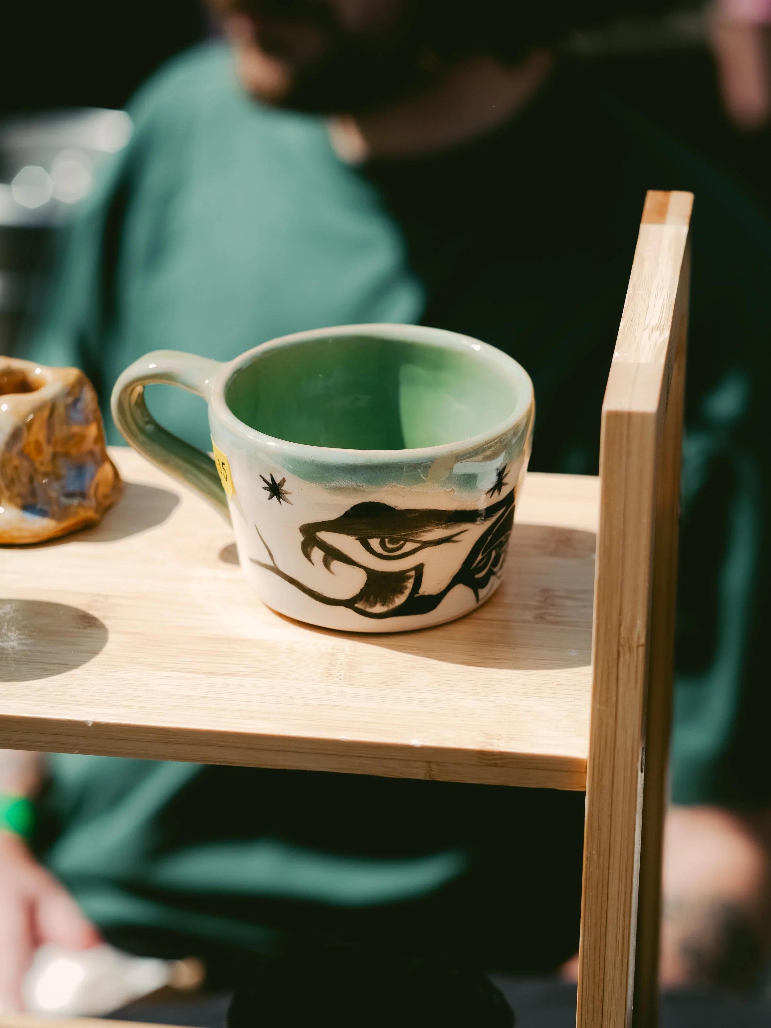Decorative ceramic mug with a green interior and black patterned design on the exterior, placed on a wooden shelf.