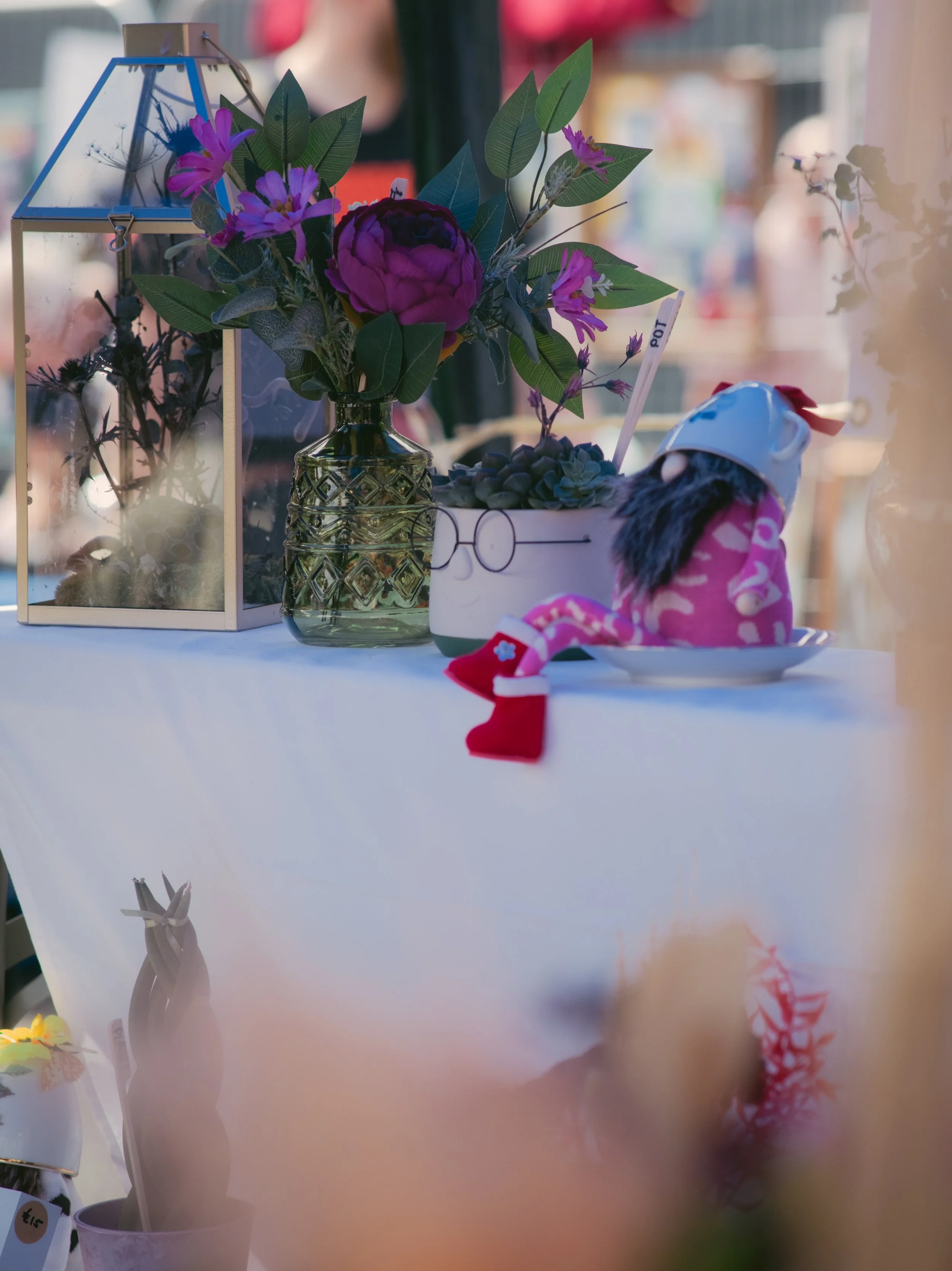 A table displays a bouquet of purple and pink flowers in a green glass vase, a glass lantern, a small pot with succulents, a pink plush toy with red boots, and a decorative white and green container with a pair of glasses painted on it.