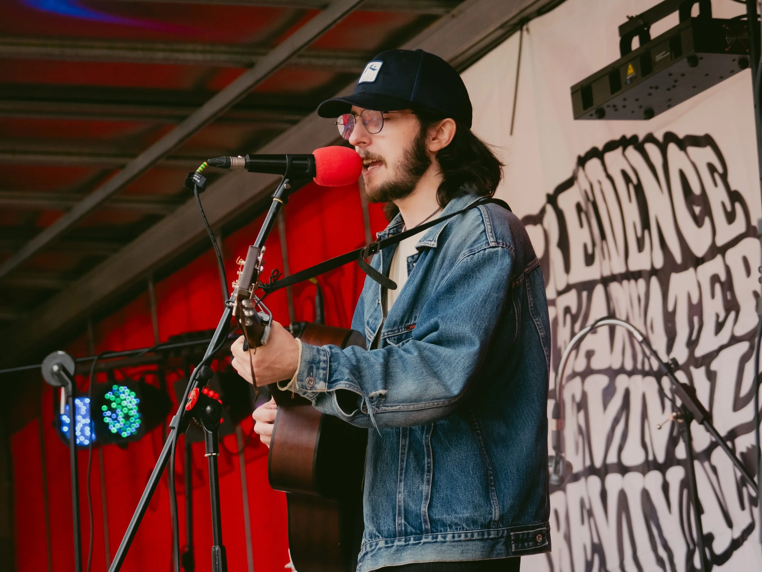 A young man with long hair, glasses, a beard, wearing a denim jacket and a black baseball cap, is singing into a microphone while playing an acoustic guitar on stage. There is a backdrop with partial text 'ENDORSE' and colored stage lights.