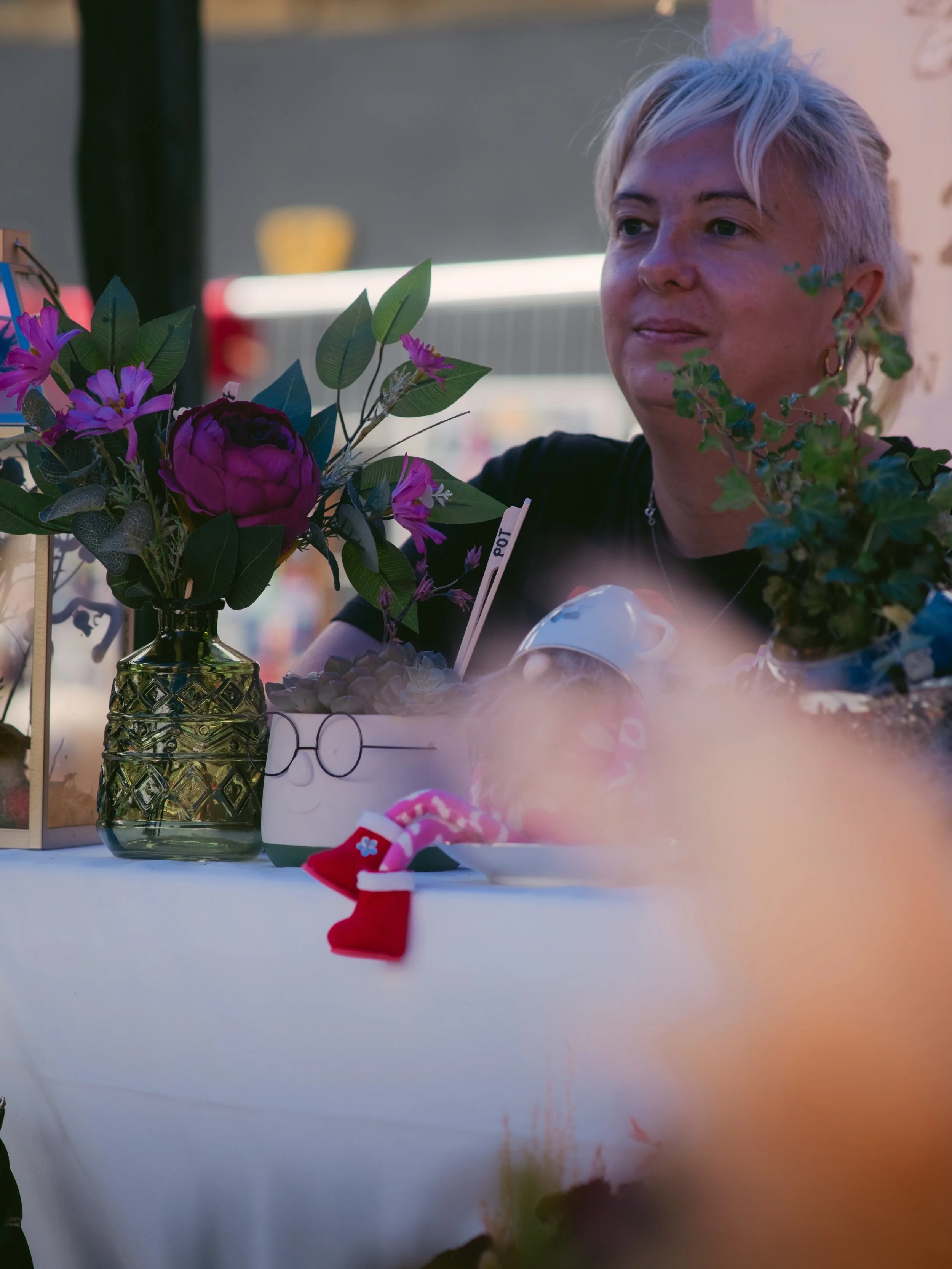 A woman with short, platinum blonde hair sits at a table decorated with flowers and small holiday-themed accessories, smiling and looking off to the side.