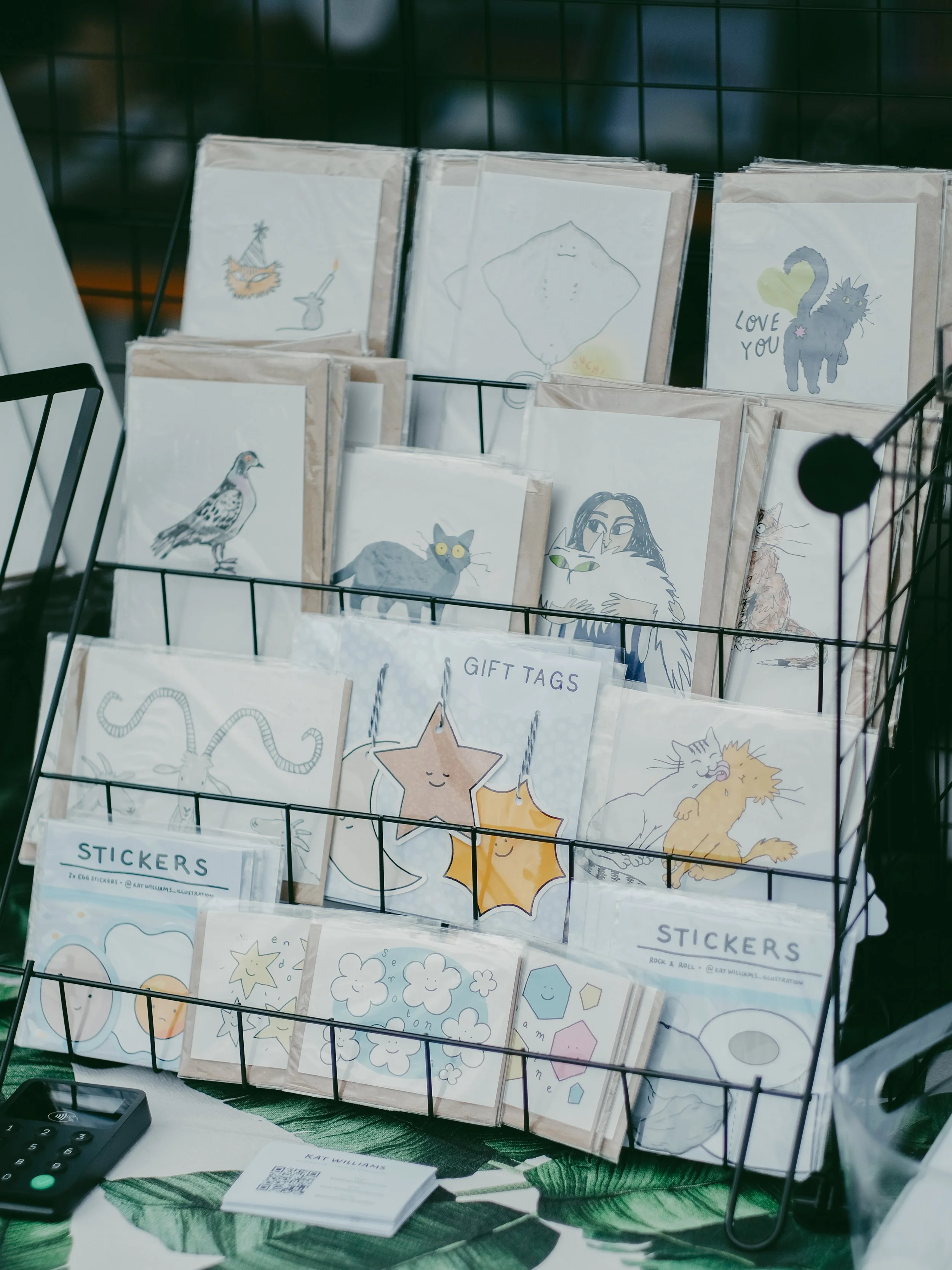 Display of greeting cards and stickers featuring hand-drawn illustrations of cats, humans, and abstract designs in a wire basket at a craft market.