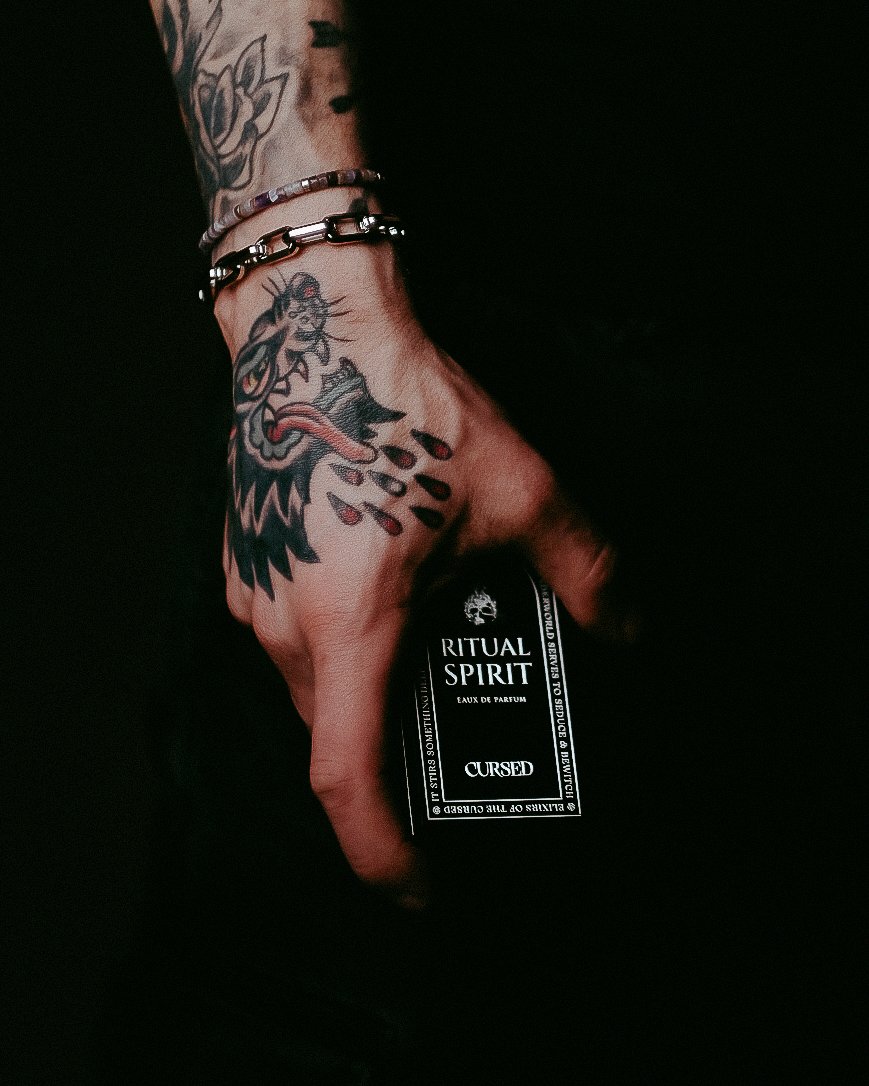 A tattooed hand holding a black bottle with a label that reads 'Ritual Spirit' and 'Cursed'. The hand has two bracelets and features a dragon tattoo on the wrist and forearm.