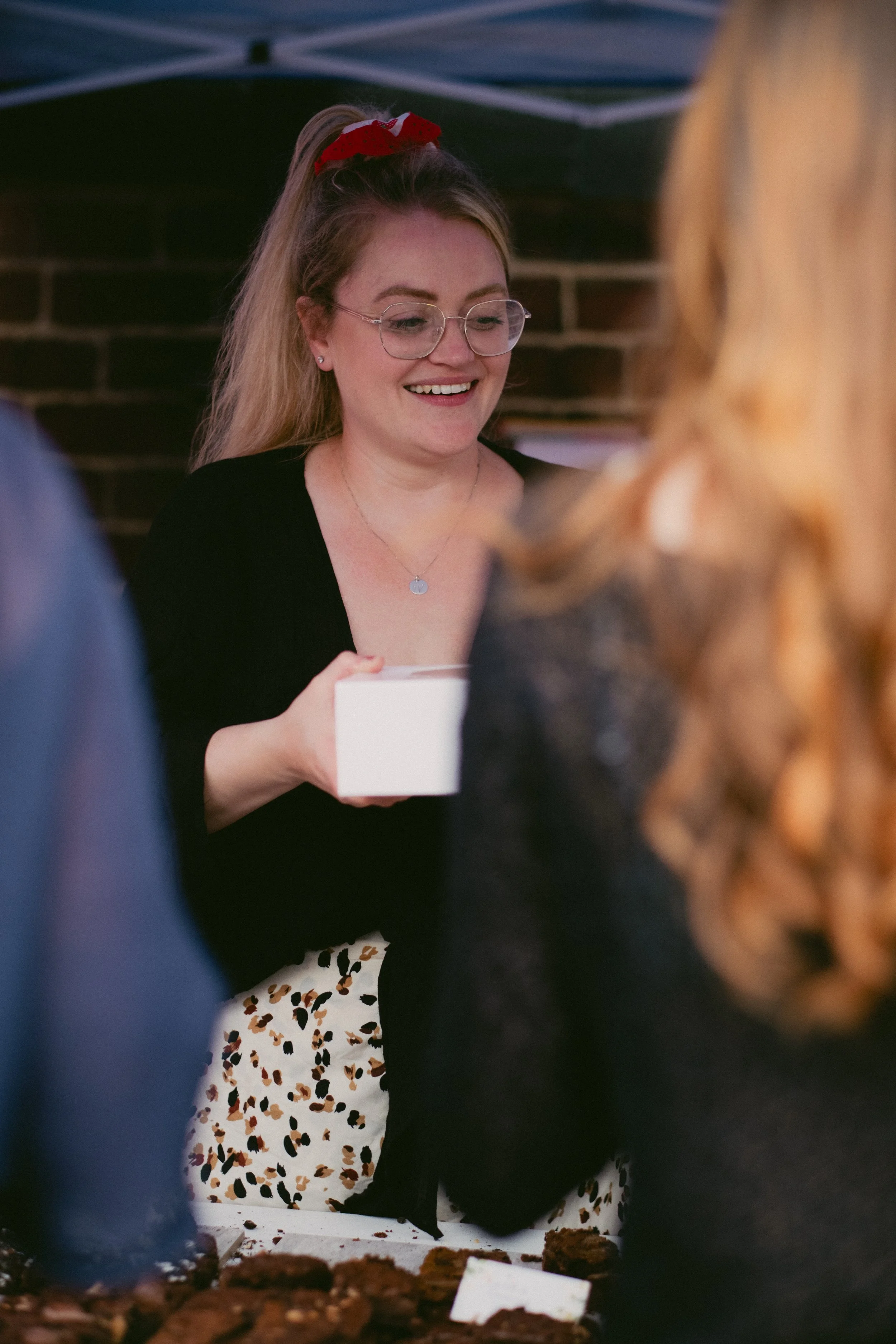 A woman with glasses and a red hair bow smiling at a gathering, holding a small white box, with other people around and a table with baked goods in front of her.