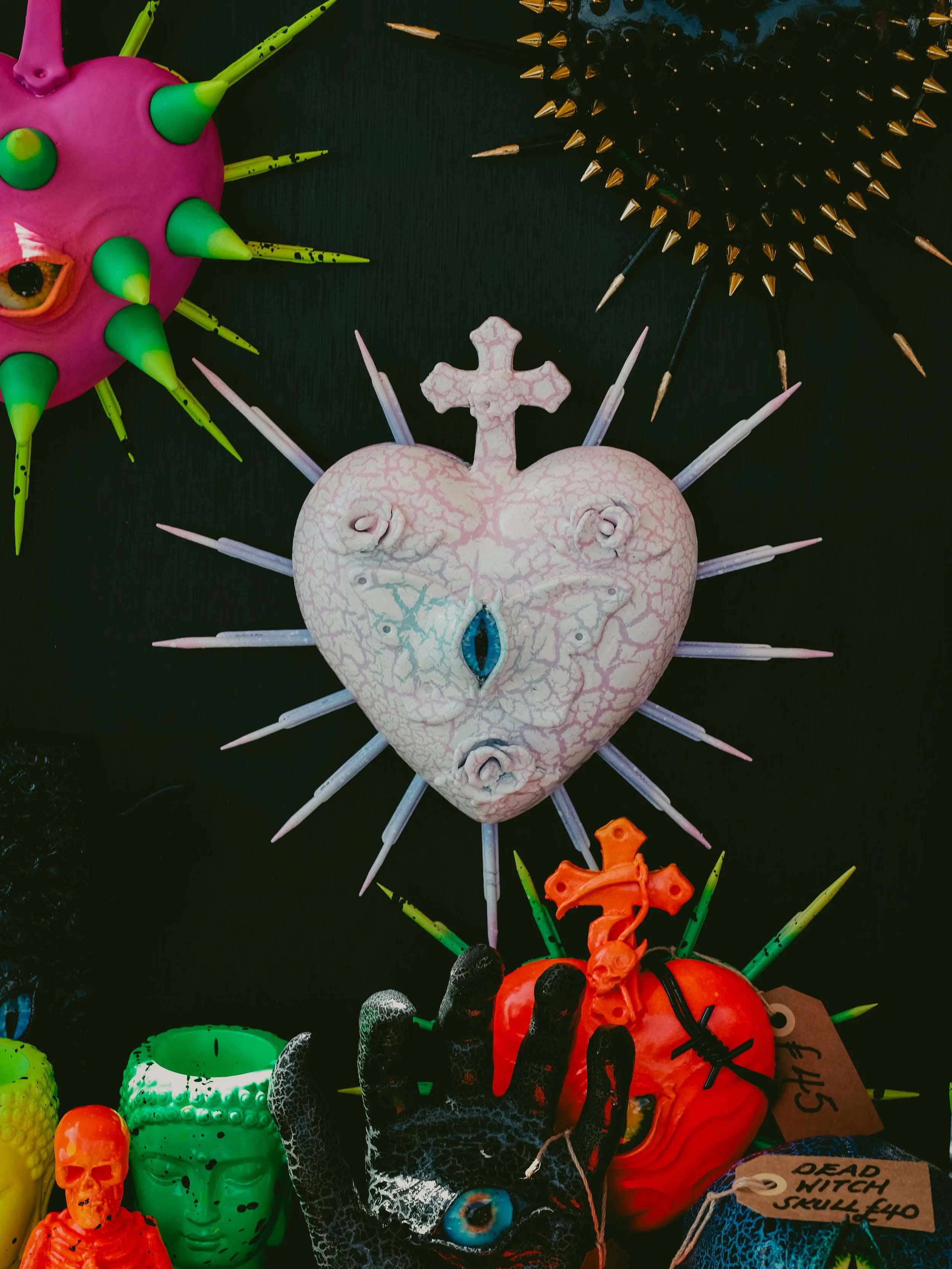Collection of Halloween decorations including a pink face with green spikes, a black spiked ball, a heart with a cross on top and an eye in the center, a small orange skull, a green face, a black hand with an eye, and a large orange pumpkin with gree