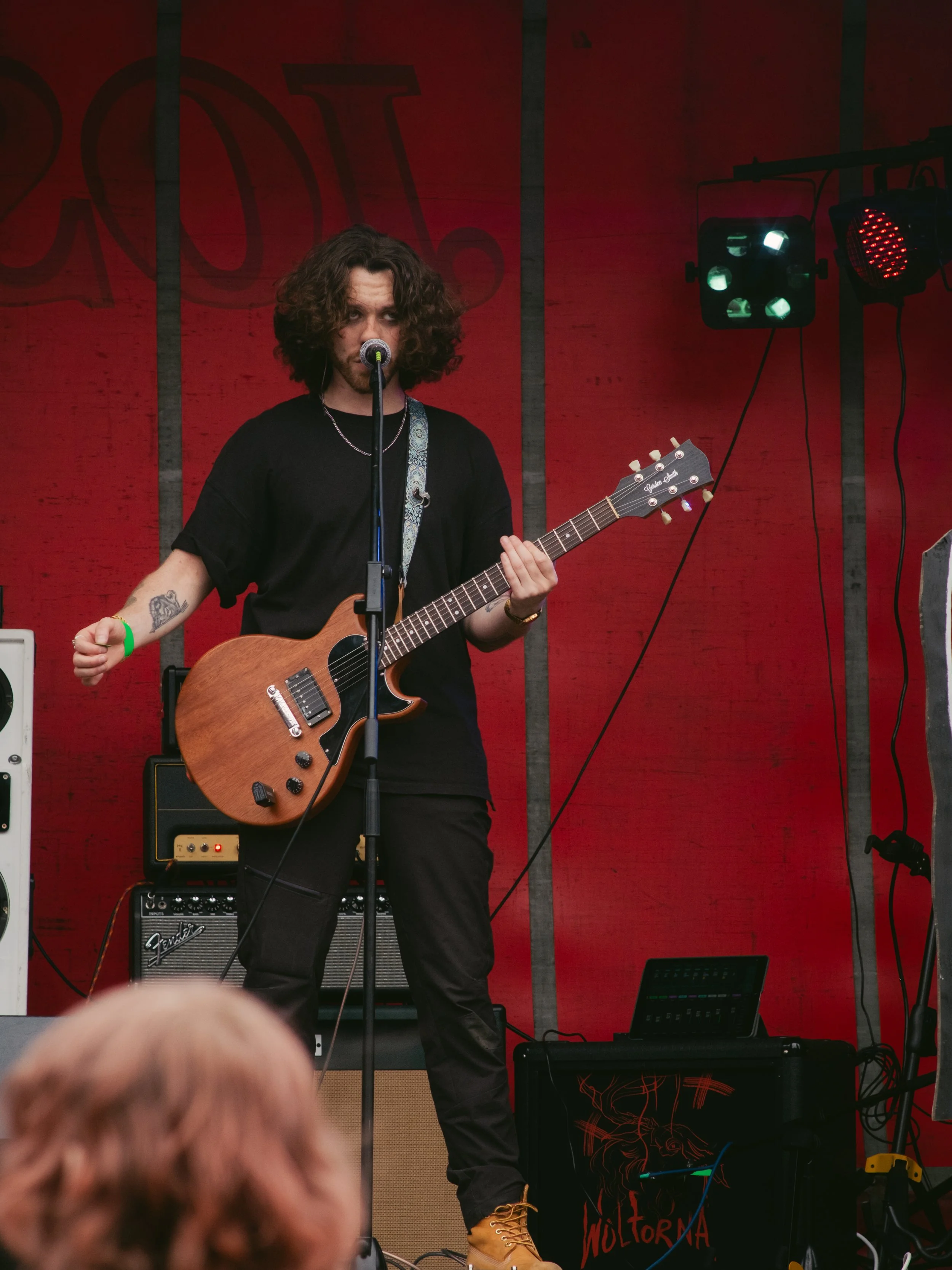 A musician with curly hair wearing a black shirt and brown shoes playing an electric guitar on stage with red backdrop and stage lighting.