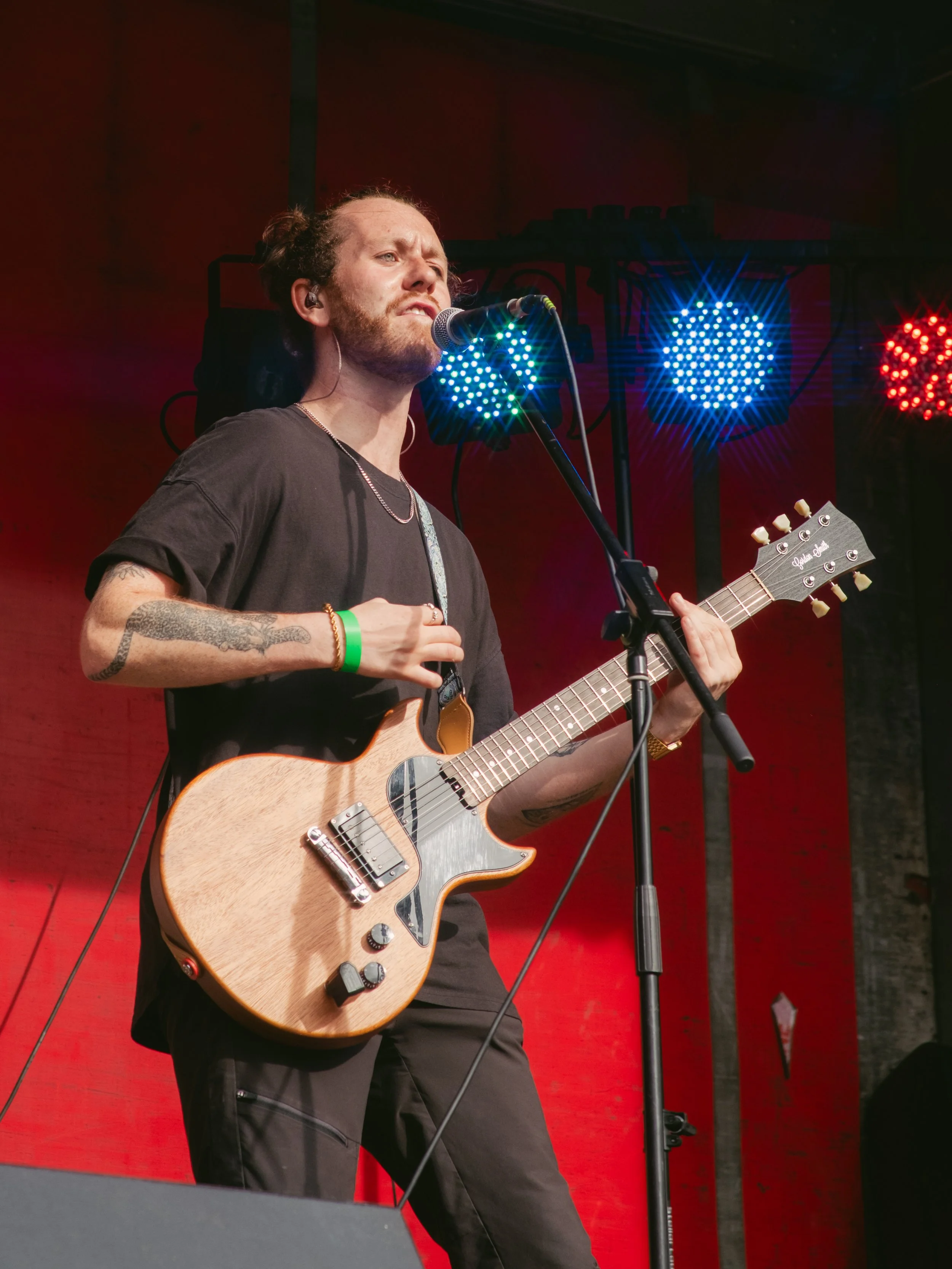 A man with a beard and tattoos playing an electric guitar and singing into a microphone on stage with colorful stage lights behind him.