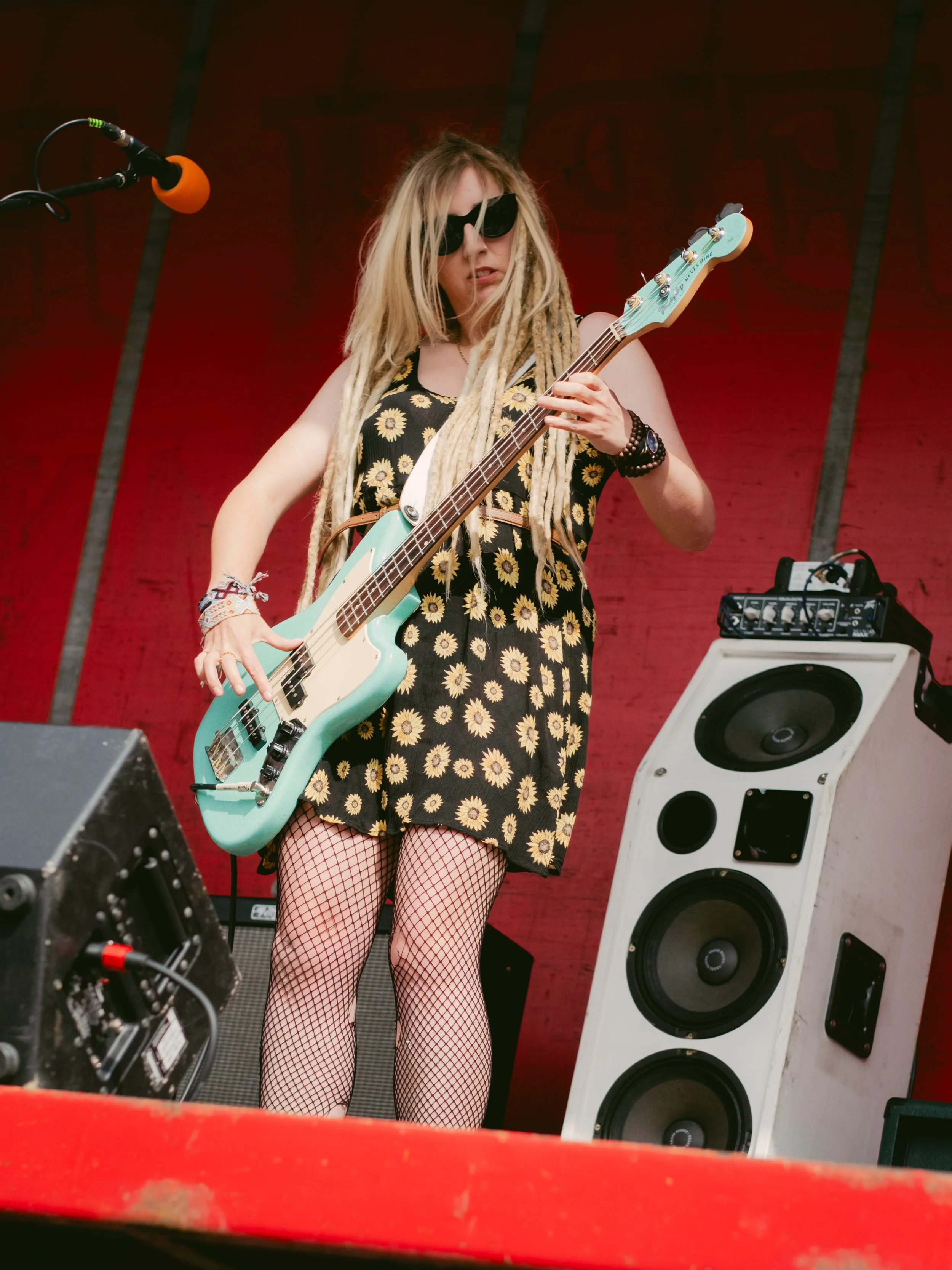 A woman with blonde dreadlocks wearing sunglasses, a sunflower dress, and fishnet stockings plays a turquoise bass guitar on stage with a red backdrop and musical equipment.