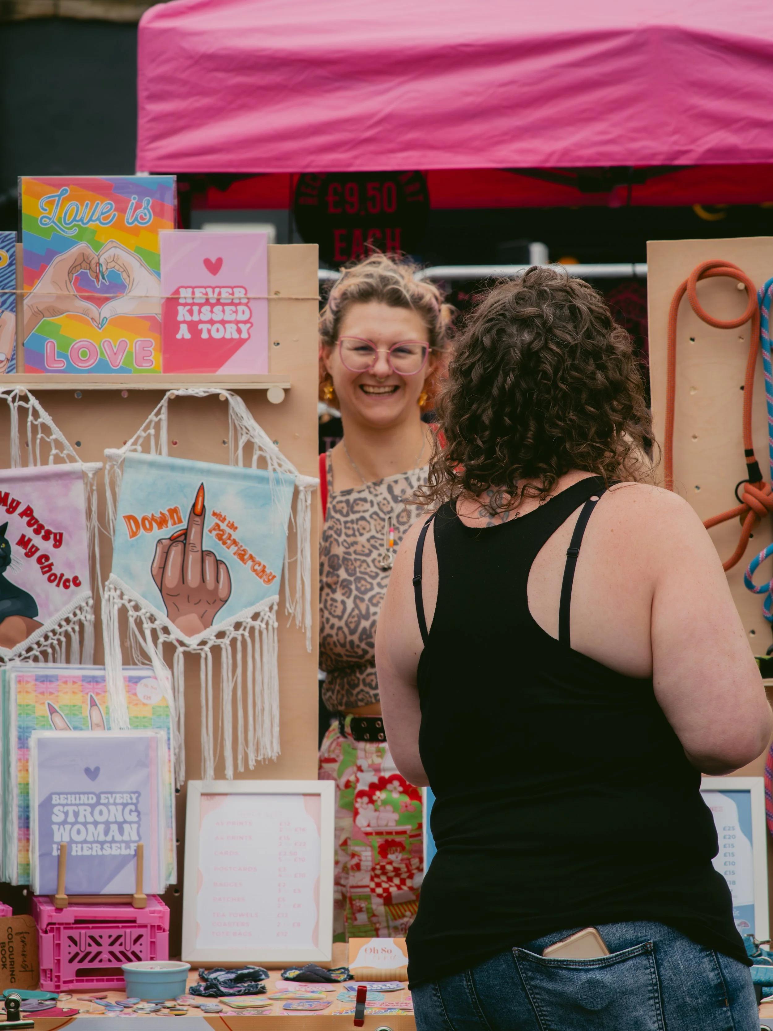 A woman with curly hair wearing a black tank top and jeans talking to a vendor at an outdoor market stall decorated with colorful signs and handmade crafts, with another woman smiling behind the stall.
