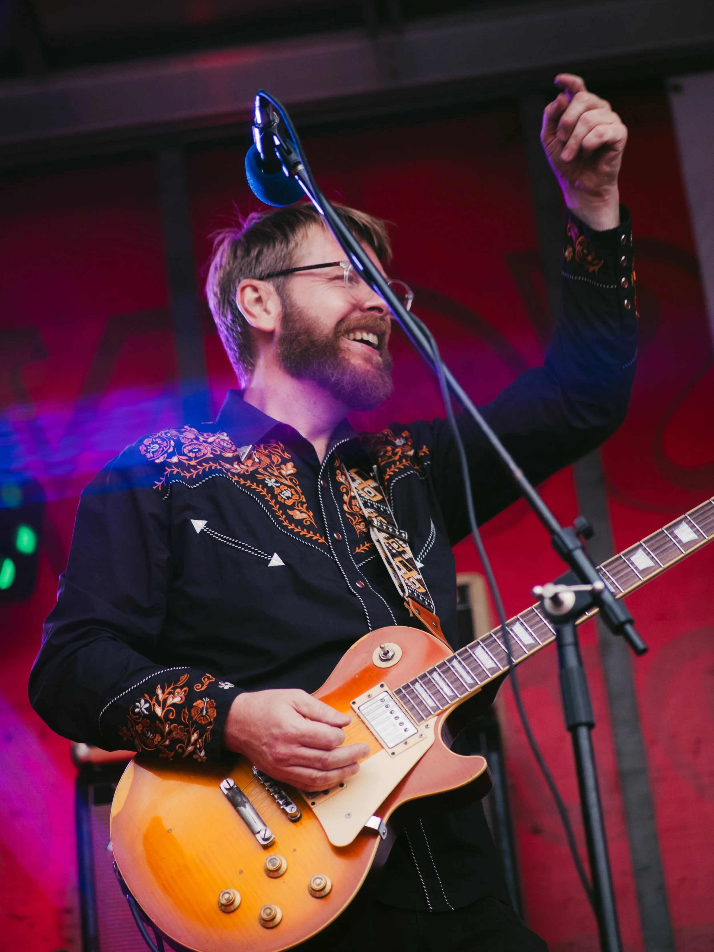 A man with glasses and a beard playing an electric guitar, smiling, with one arm raised in front of a red wall background during a live music performance.