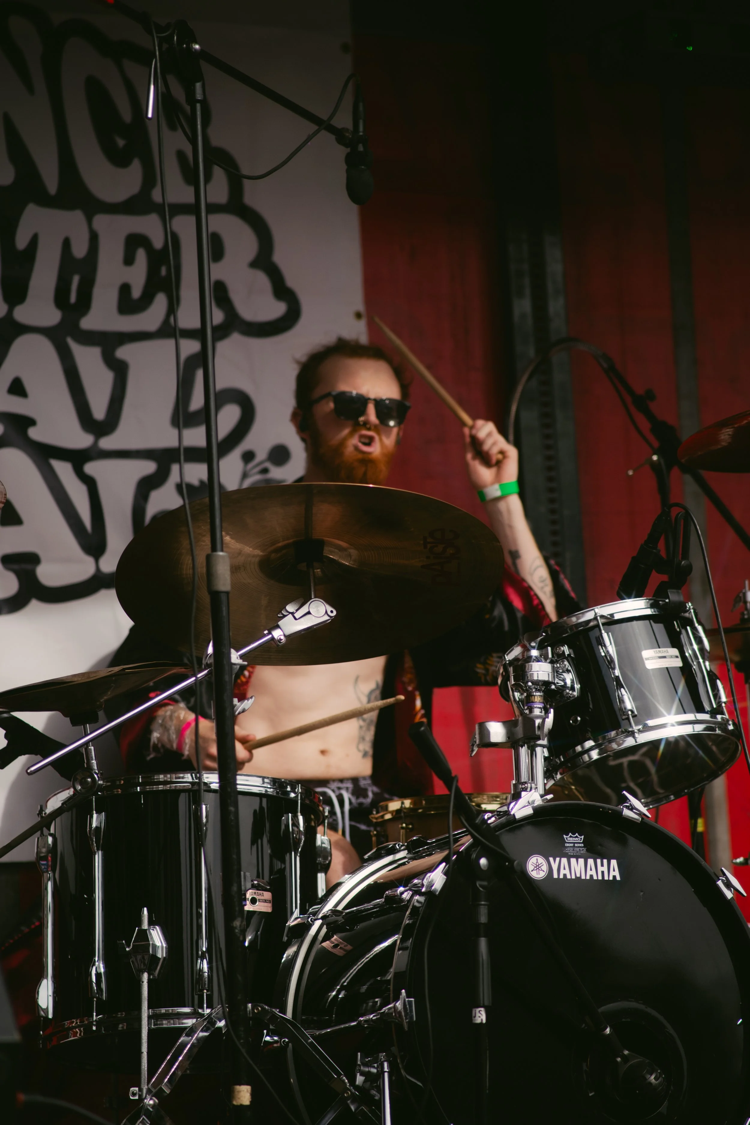 A man with sunglasses and a beard playing a drum set on stage during a performance.