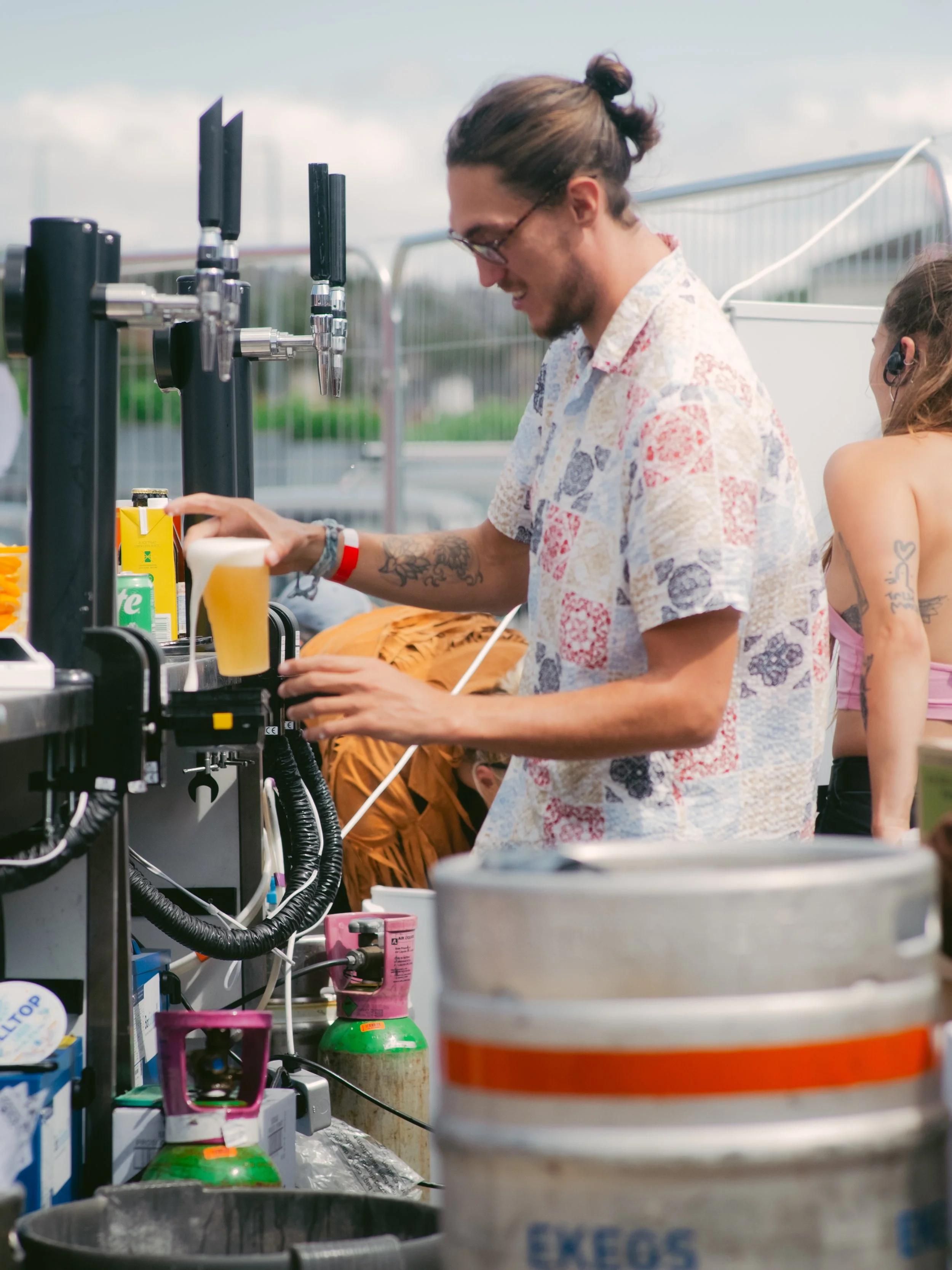 A man with glasses and a man bun pours a draft beer at an outdoor bar or festival stand, with a woman in the background and propane tanks and drinks nearby.