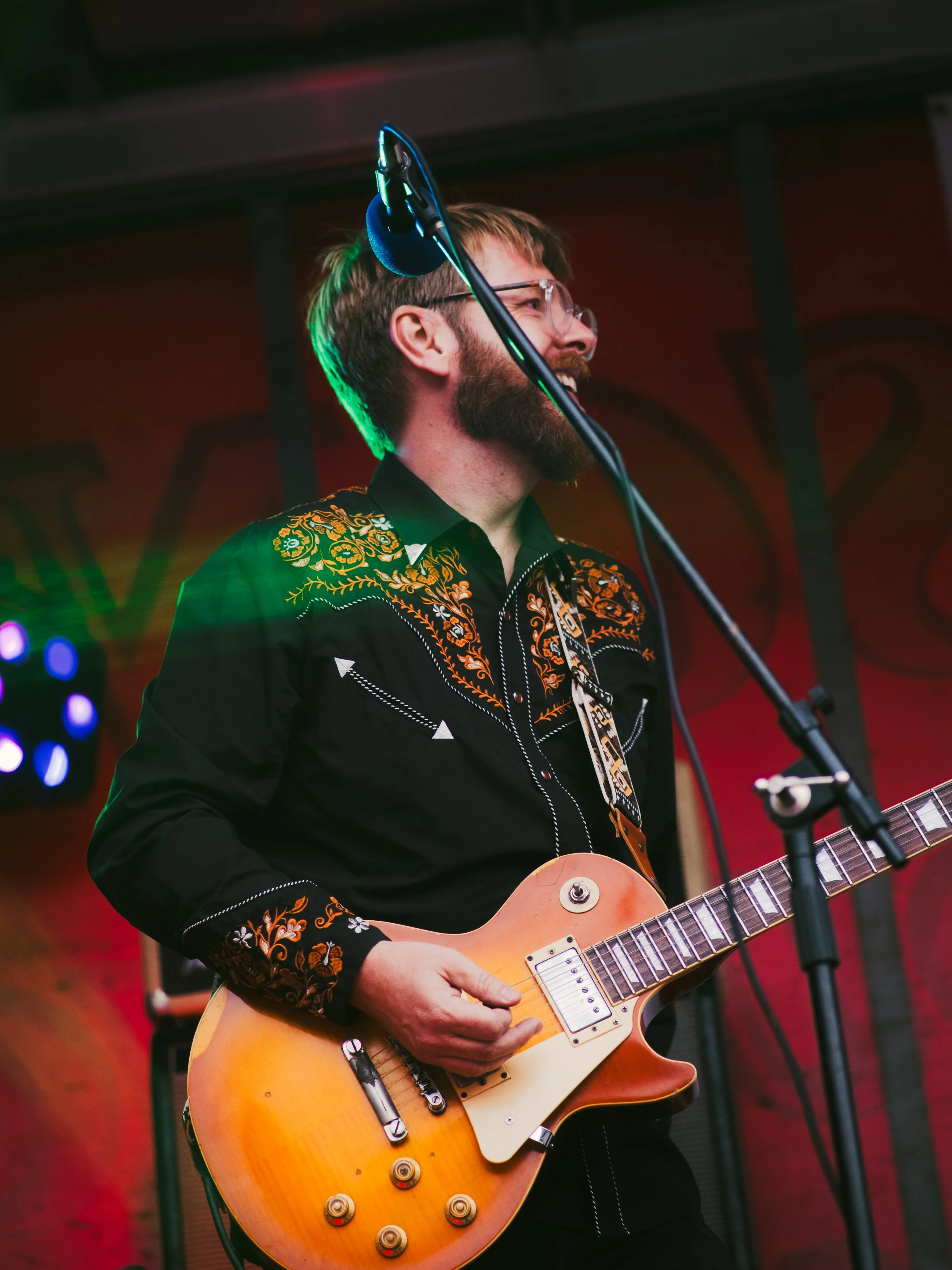 A man with glasses and a beard playing an orange electric guitar on stage, wearing a black western shirt with orange floral embroidery.
