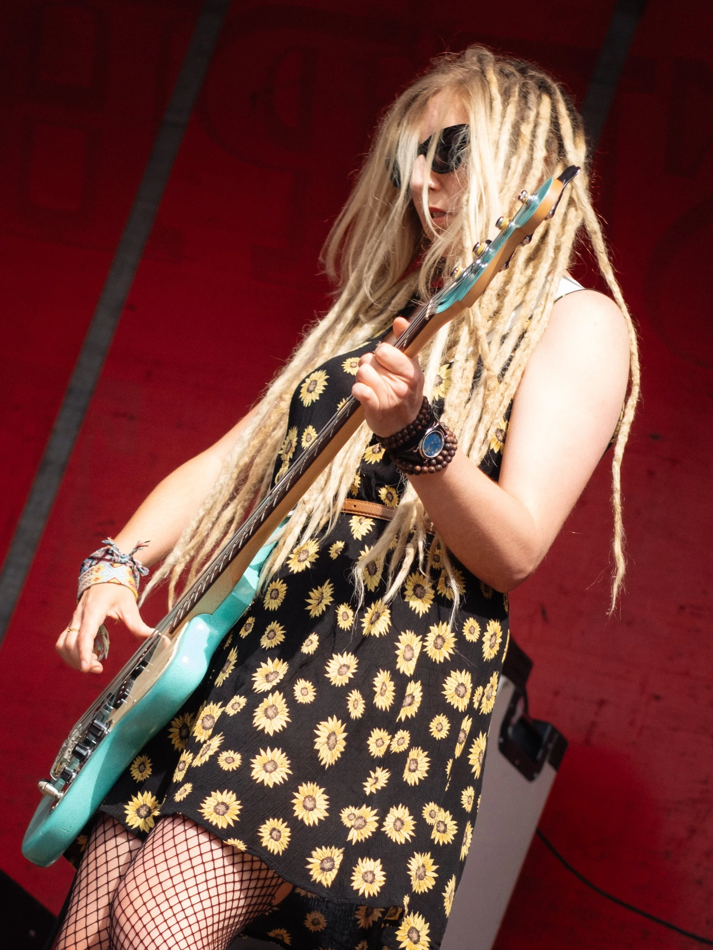 A woman with long, blonde dreadlocks wearing sunglasses, a black dress with a sunflower pattern, fishnet stockings, and various bracelets, playing an electric guitar.