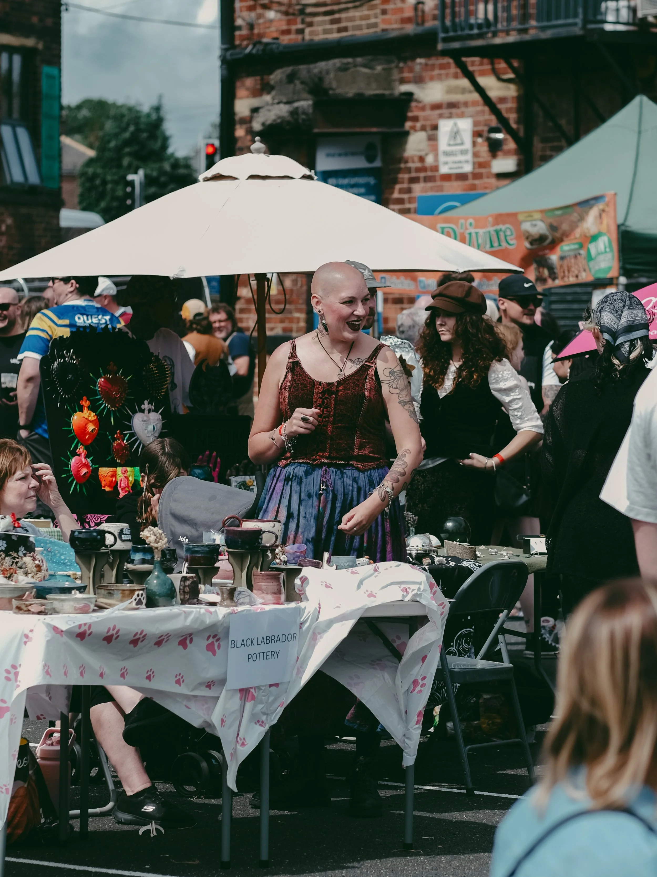 A woman with tattoos, a bald head, and a dress with a corset is smiling and talking to people at a marketplace stall under a white umbrella. The stall has pottery items and a sign that reads "Black Labrador Pottery." There are many people around, som