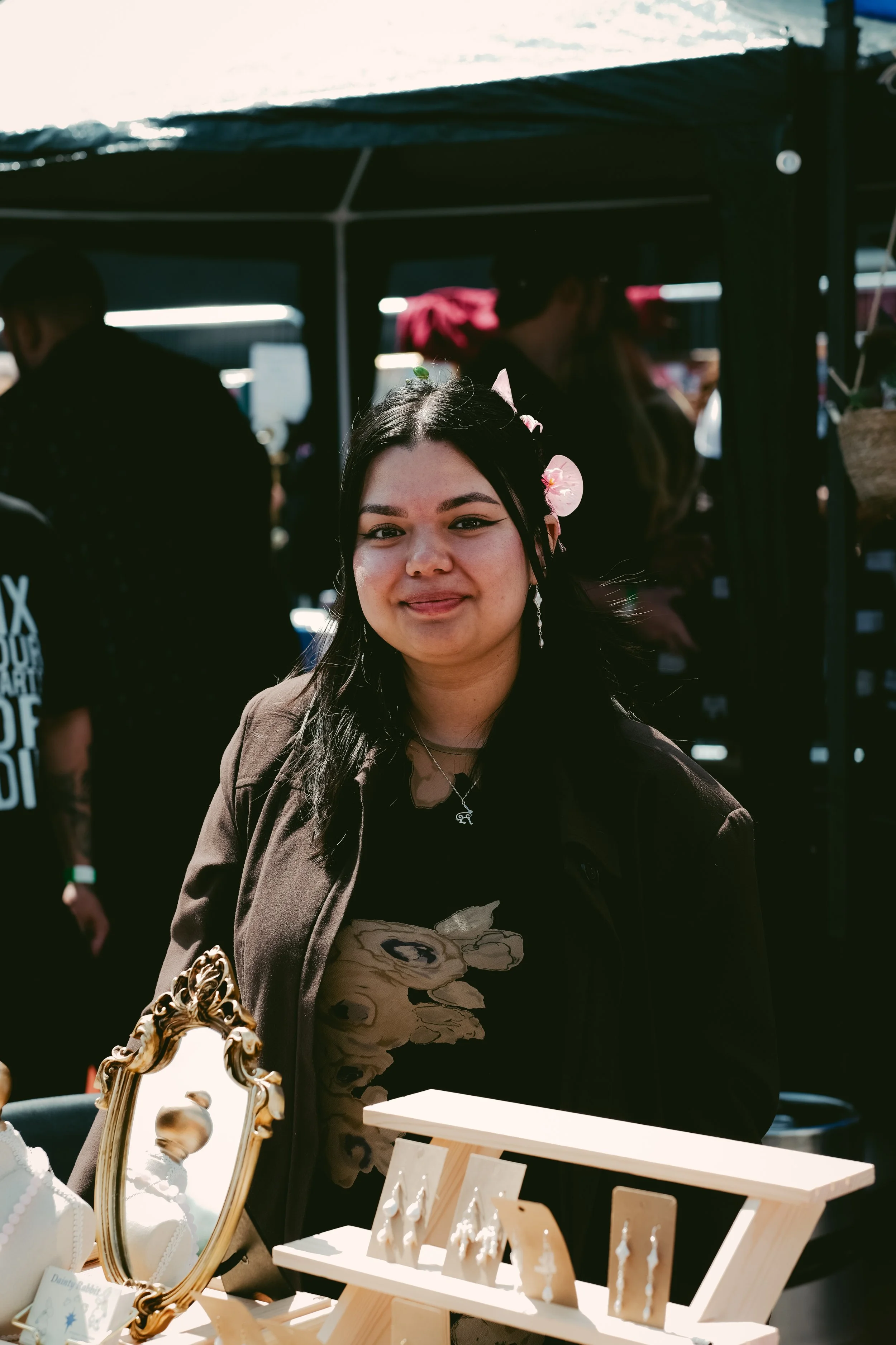 A woman with dark hair, floral hair accessory, and earrings standing at a market stall with jewelry.