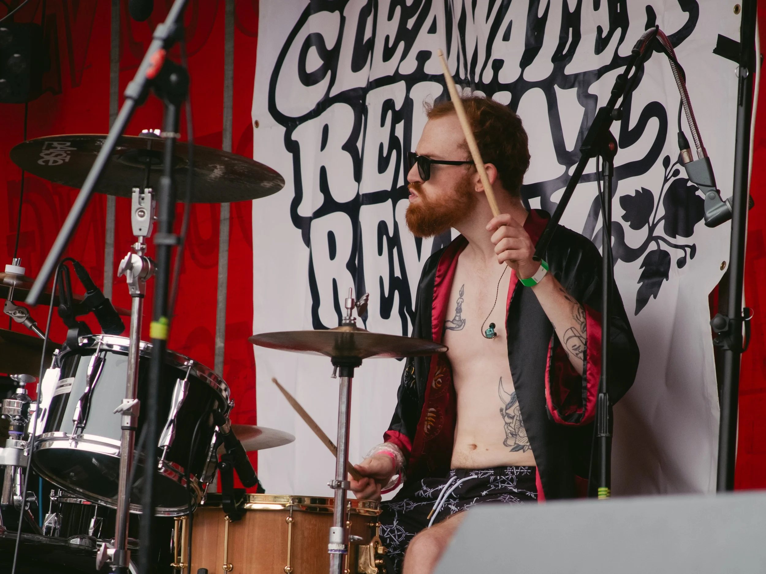 A man with tattoos, glasses, and a beard playing drums on stage at an outdoor event. He's shirtless with an open black and red robe, wearing headphones and holding a drumstick in each hand. There is a large banner behind him with black text on a whit