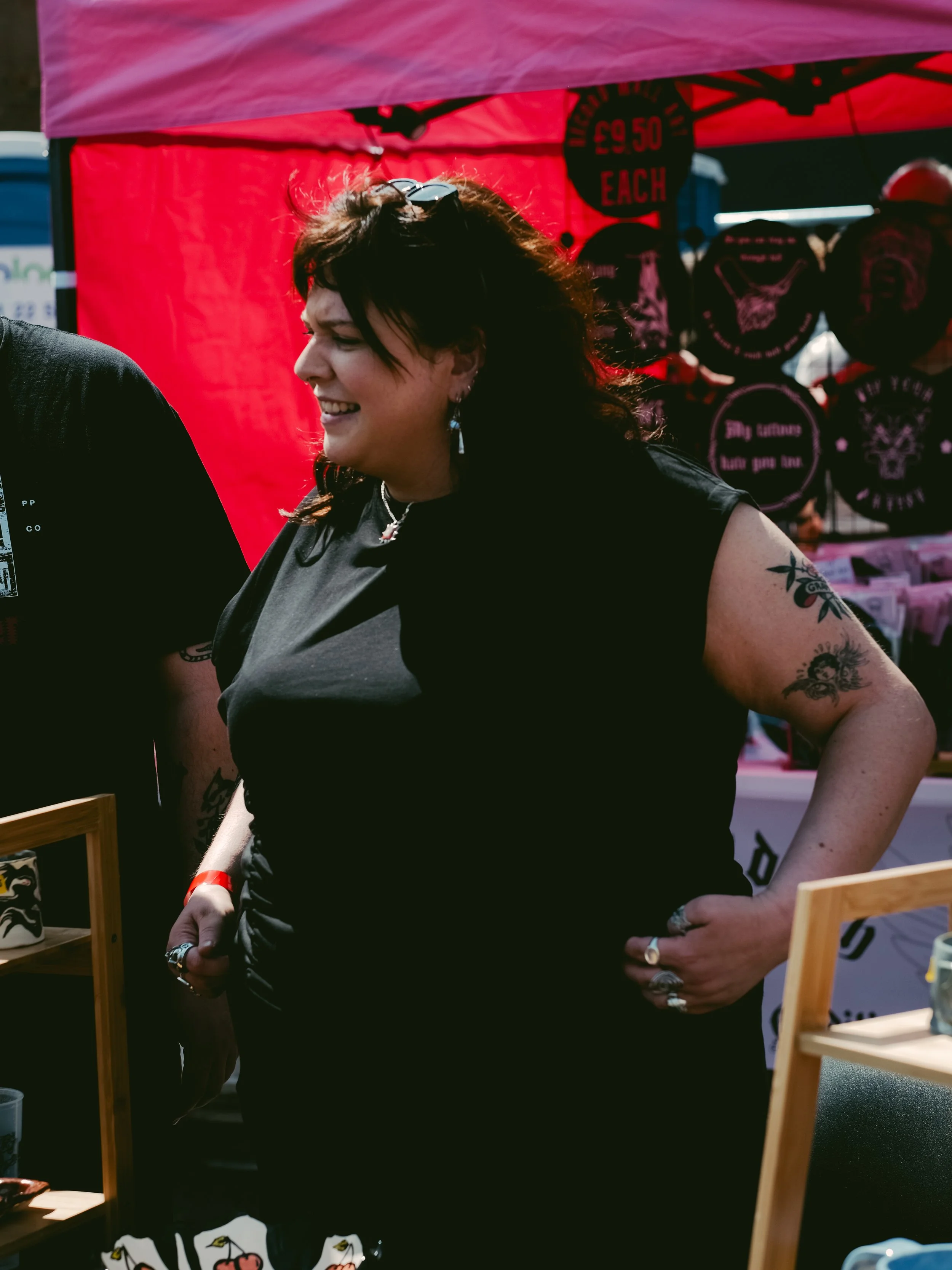 Woman with dark hair, tattoos, and rings smiling at a market stall, wearing a black shirt, in front of a red backdrop with signs for various products.
