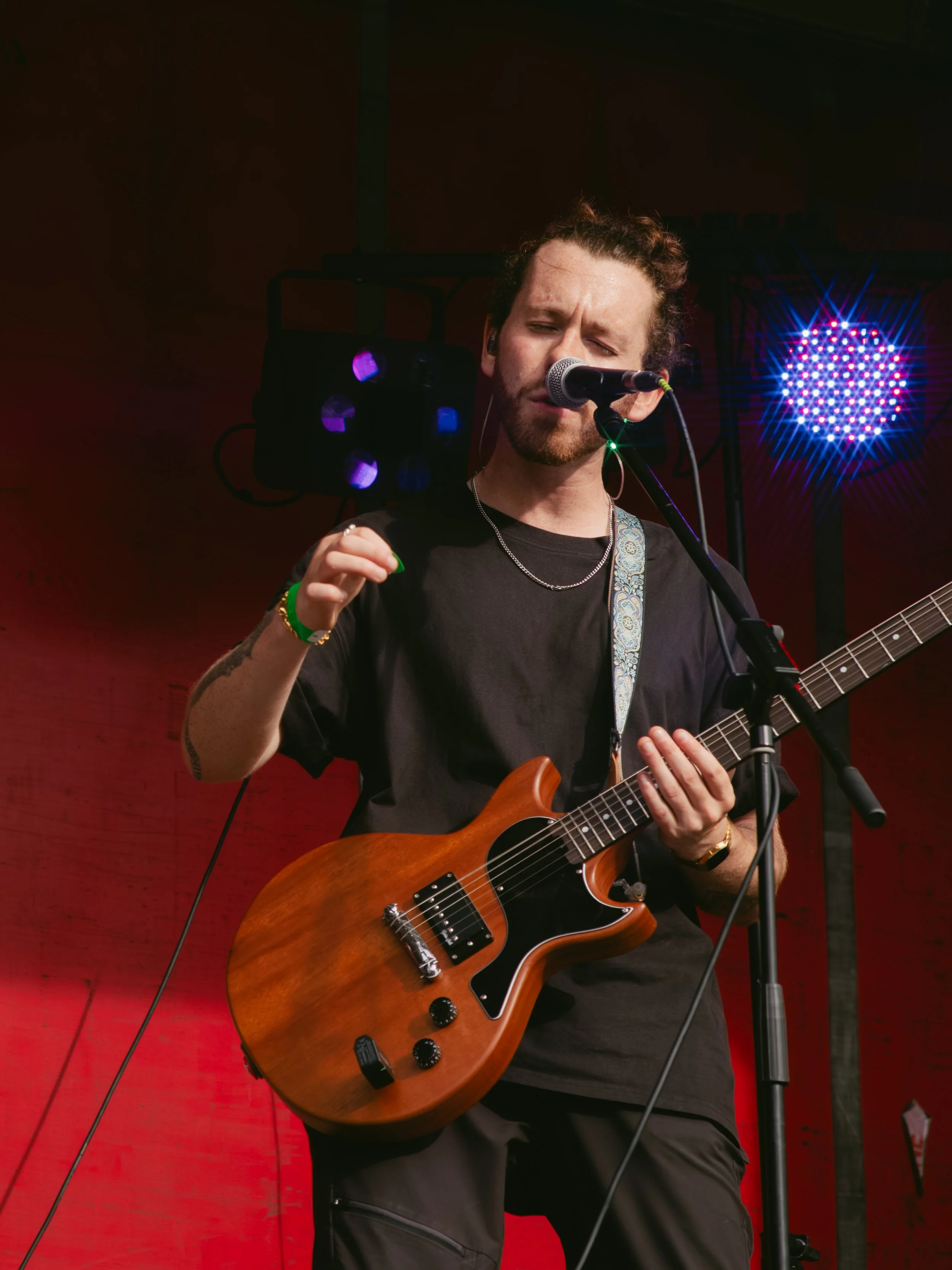 A male musician with a guitar performing on stage, singing into a microphone, with colorful stage lights in the background.