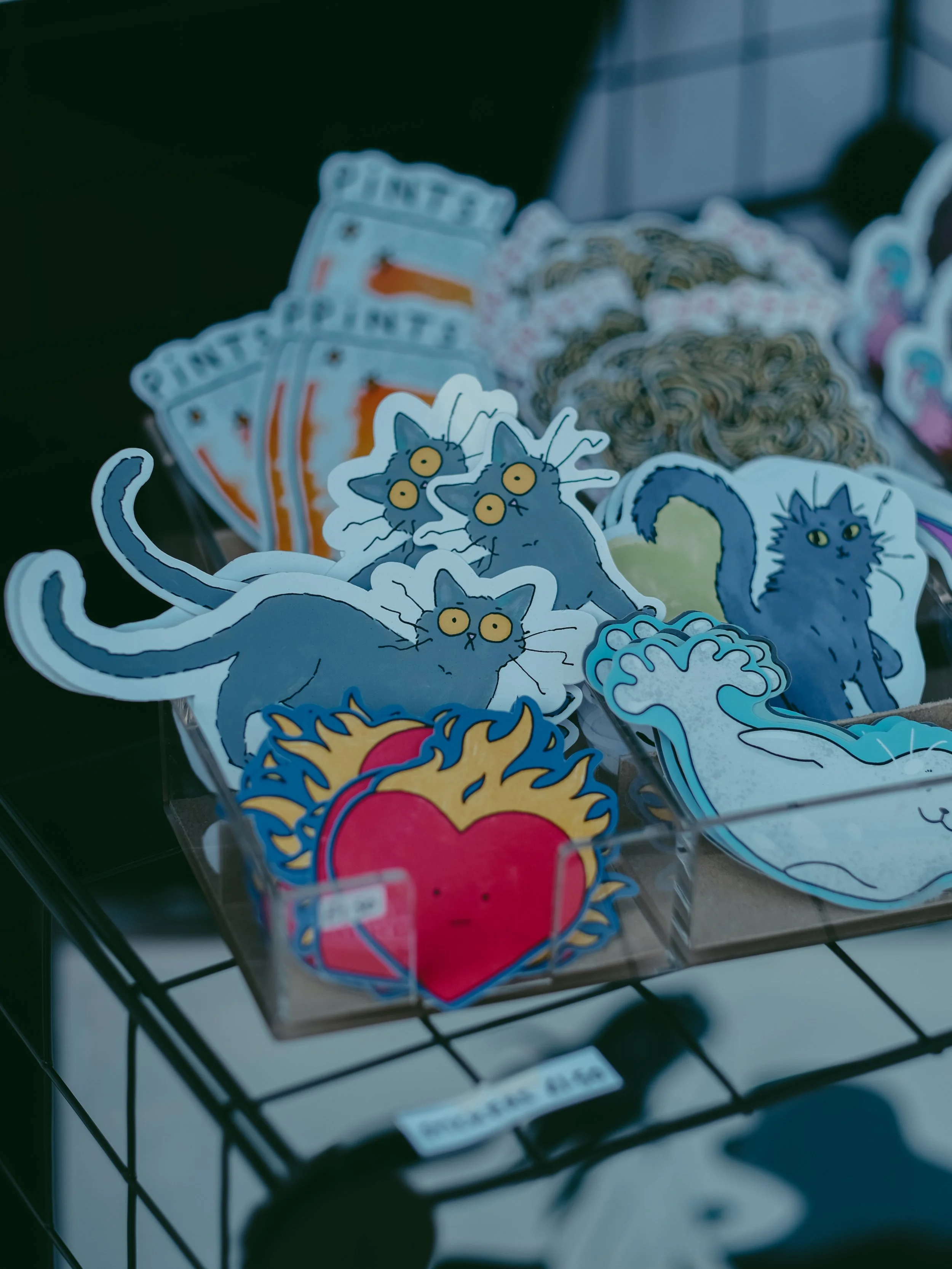 Sticker pack featuring cartoon cats, a flaming heart, and other designs on display on a black wire rack.