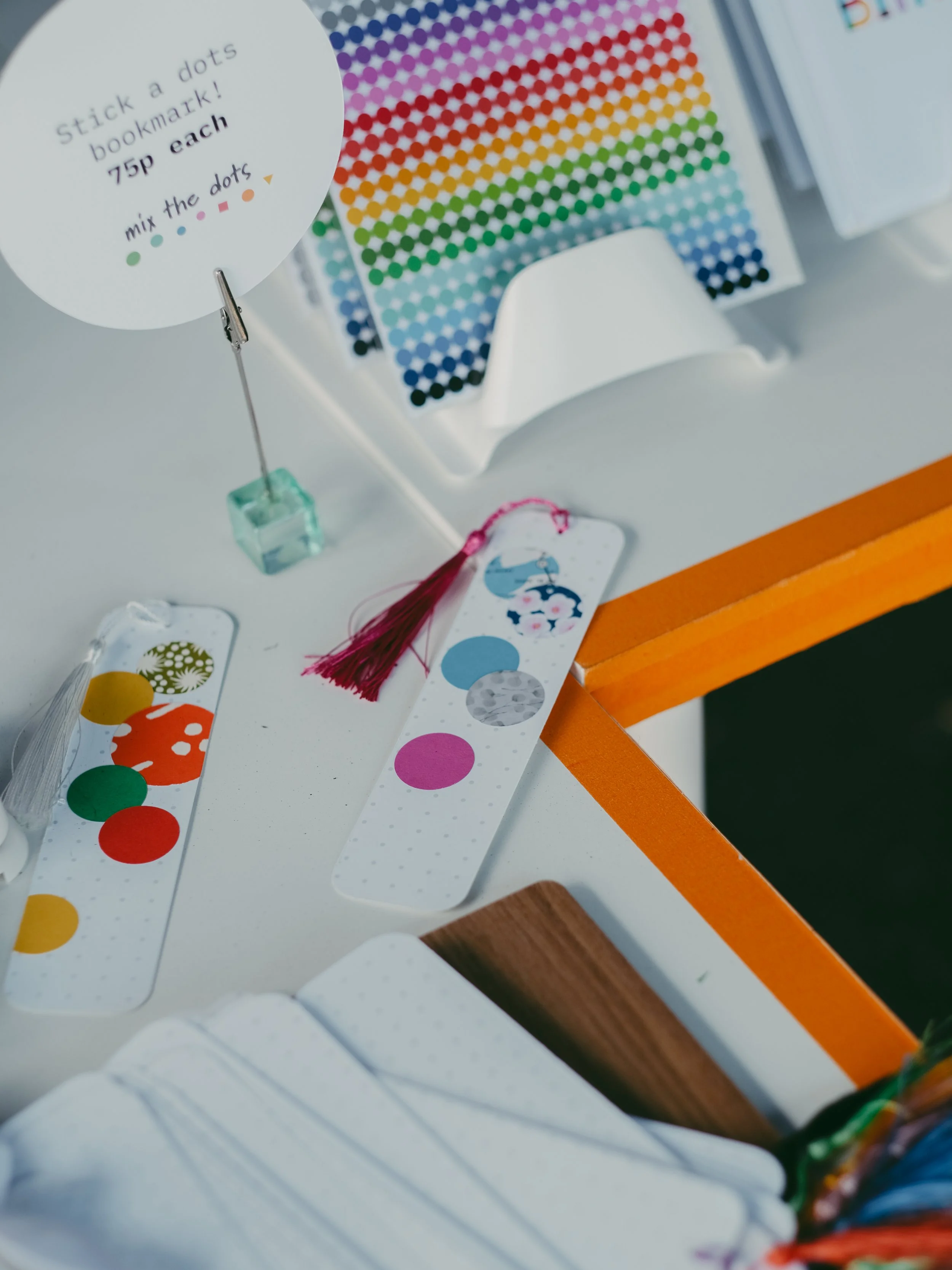 Display of colorful dot stickers, a sign with instructions to stick dots on a bookmark for 75 cents each, clothespins holding the signs, and a collection of bookmarks with various dot designs and colors.