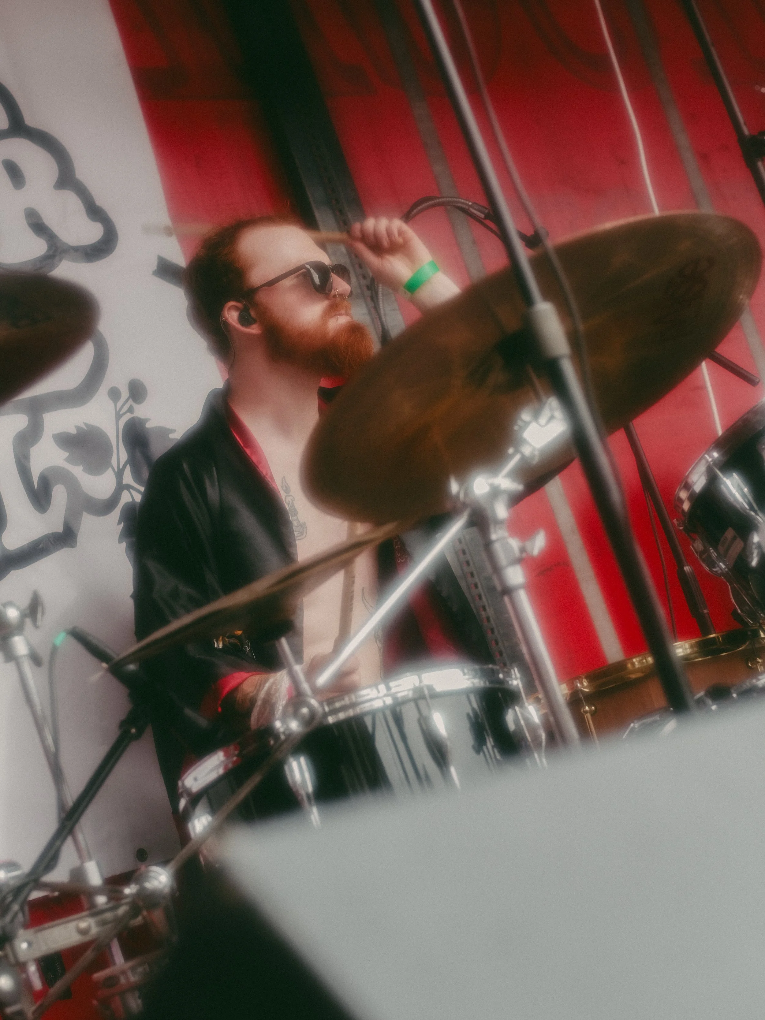 A man with red hair, a beard, sunglasses, and a black jacket playing drums on stage with a red and white background.