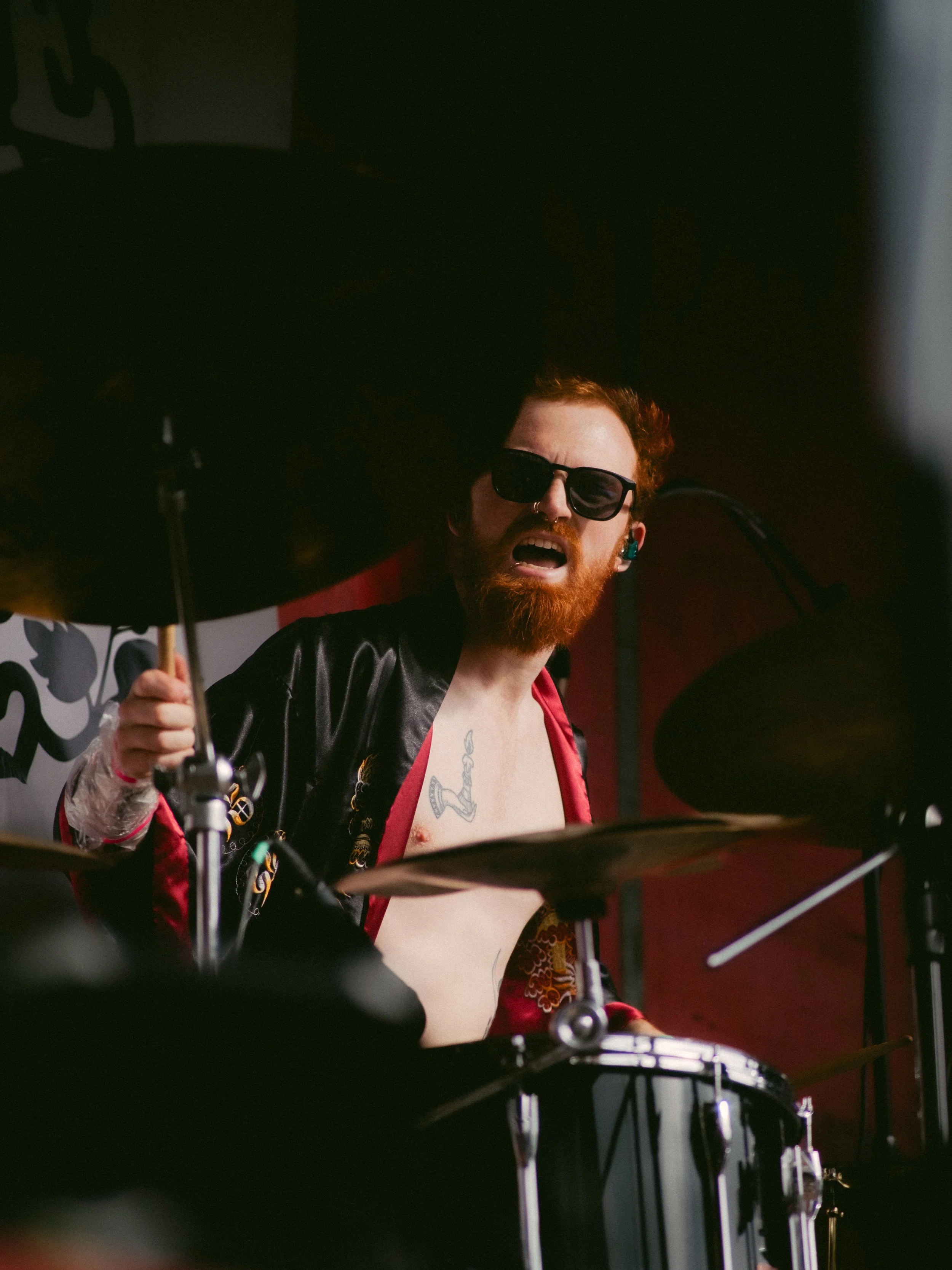 A drummer with a red beard, sunglasses, and a tattoo on his chest, performing passionately behind a drum set on stage.