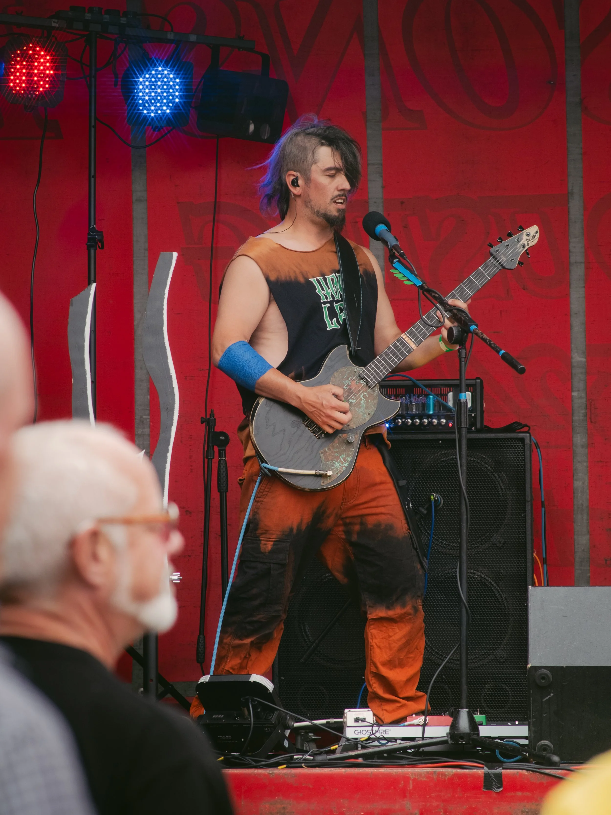 A male musician with long, colored hair, wearing a sleeveless shirt and tie-dye pants, performs on stage playing an electric guitar. He appears to be singing into a microphone in front of a red background with stage lights overhead.