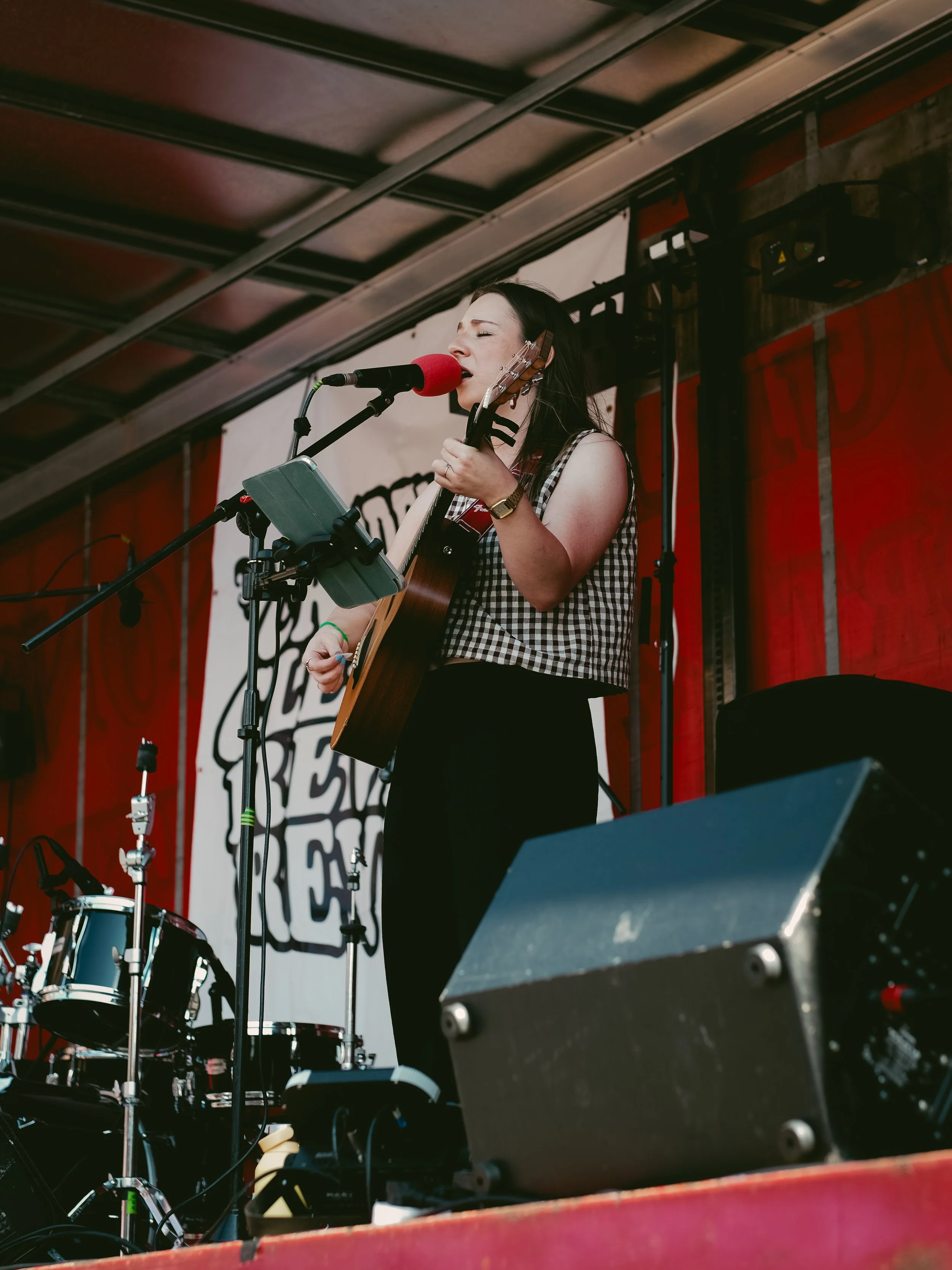 A woman singing into a microphone and playing an acoustic guitar on a stage with drums and sound equipment around her.