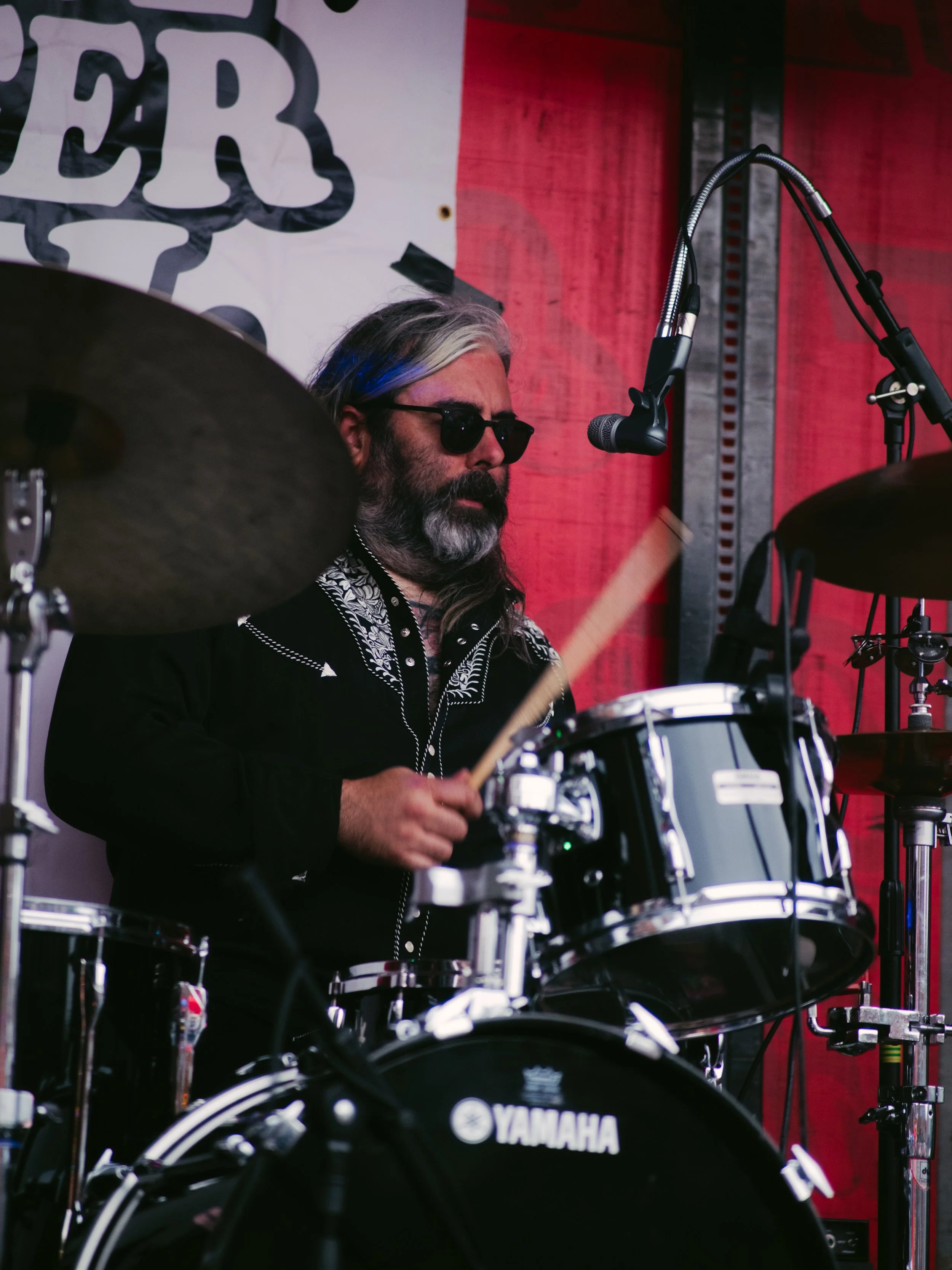 Male drummer wearing sunglasses and a black embroidered shirt playing a Yamaha drum set during a performance.