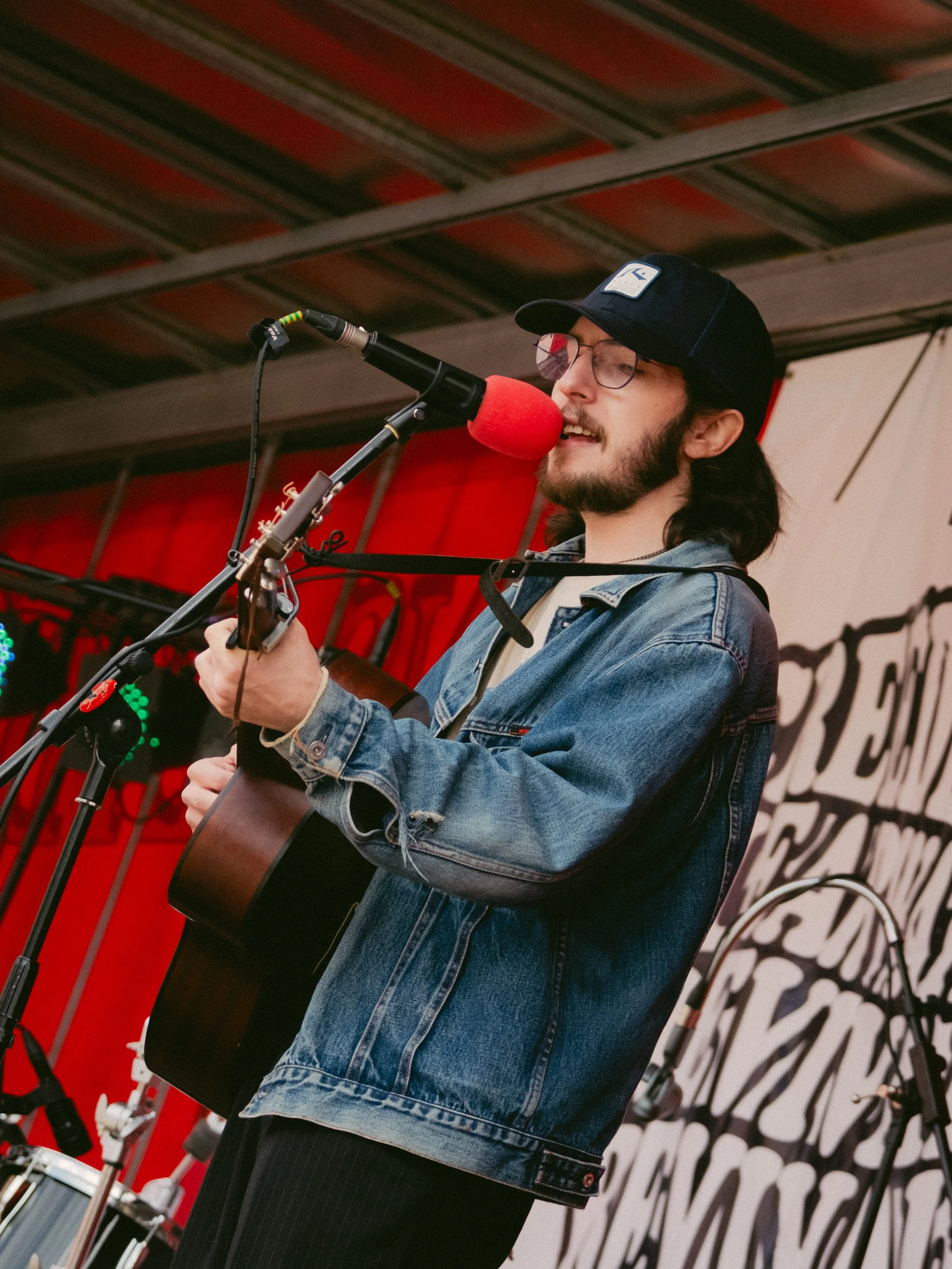 Young man with long hair, glasses, and a beard singing into a red microphone while playing an acoustic guitar on stage, wearing a denim jacket and a black cap.