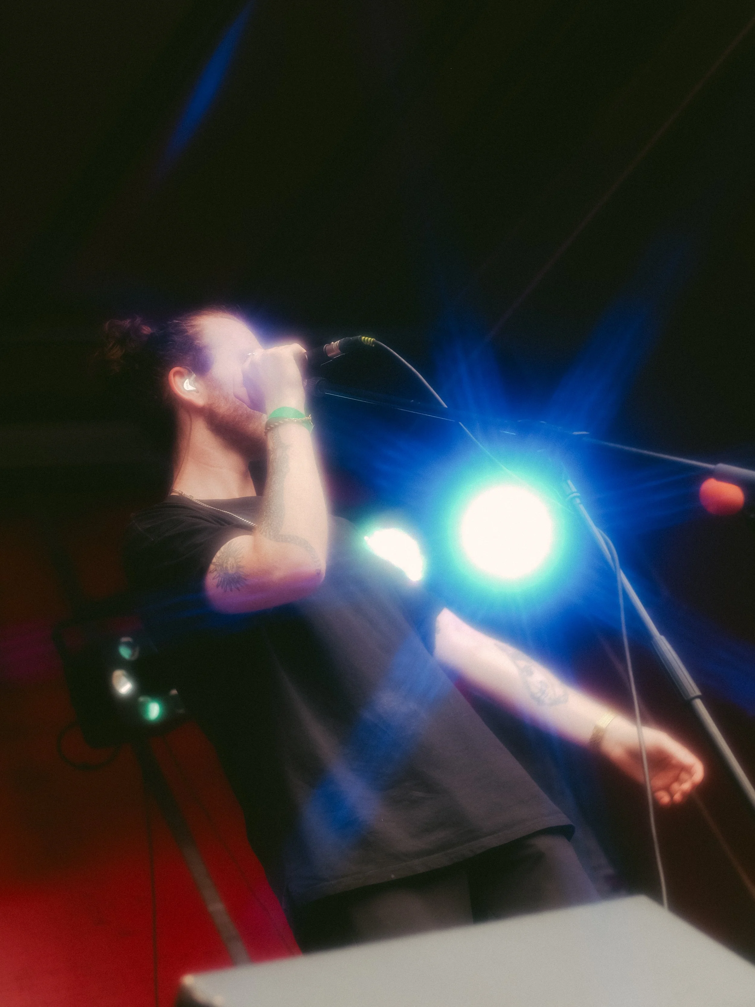A person with a bun hairstyle, tattoos, and a black shirt singing into a microphone on stage, illuminated by bright stage lights and colored lights creating a vibrant atmosphere.