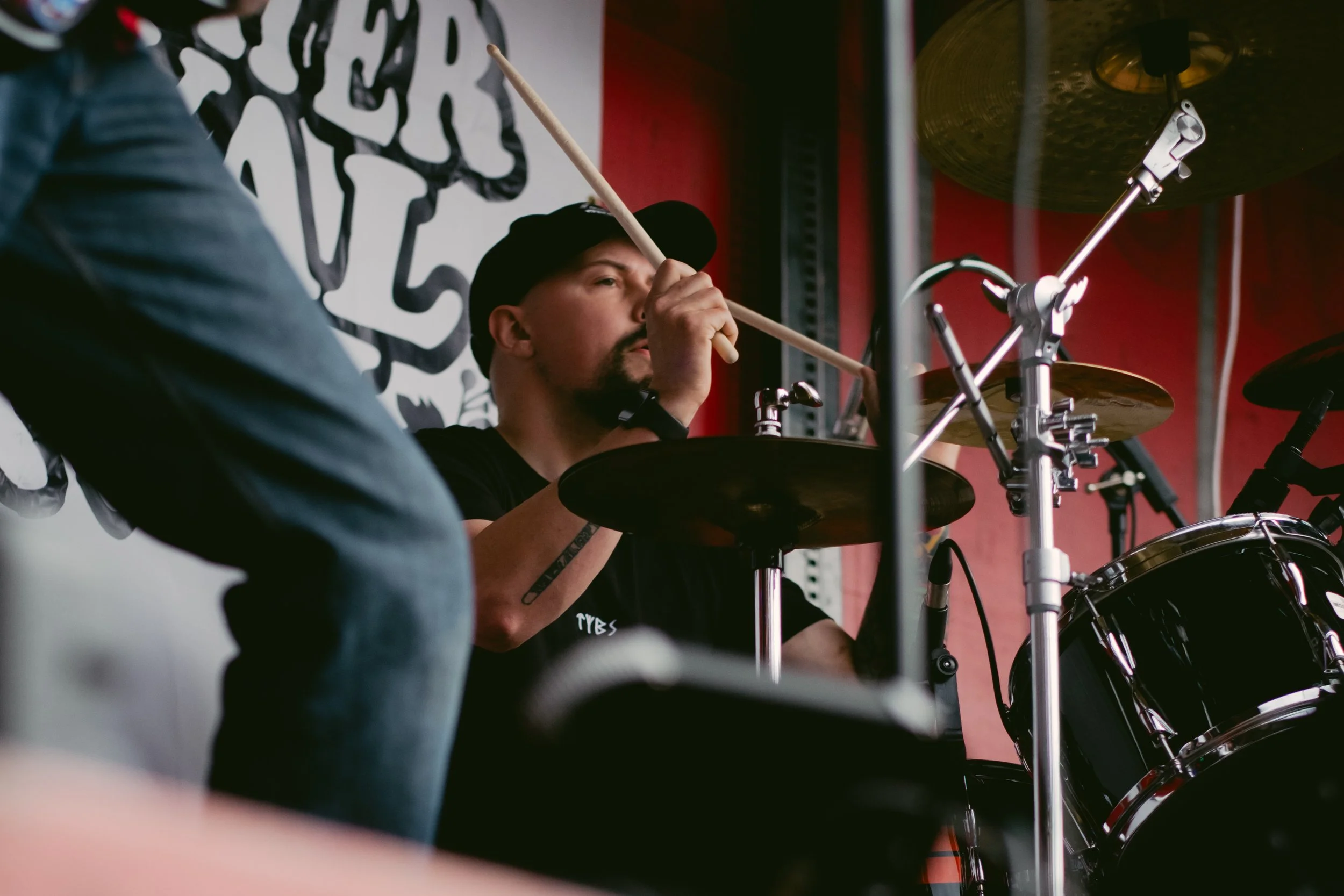 A musician playing the drums during a performance in a music venue with a red wall and black wall art.