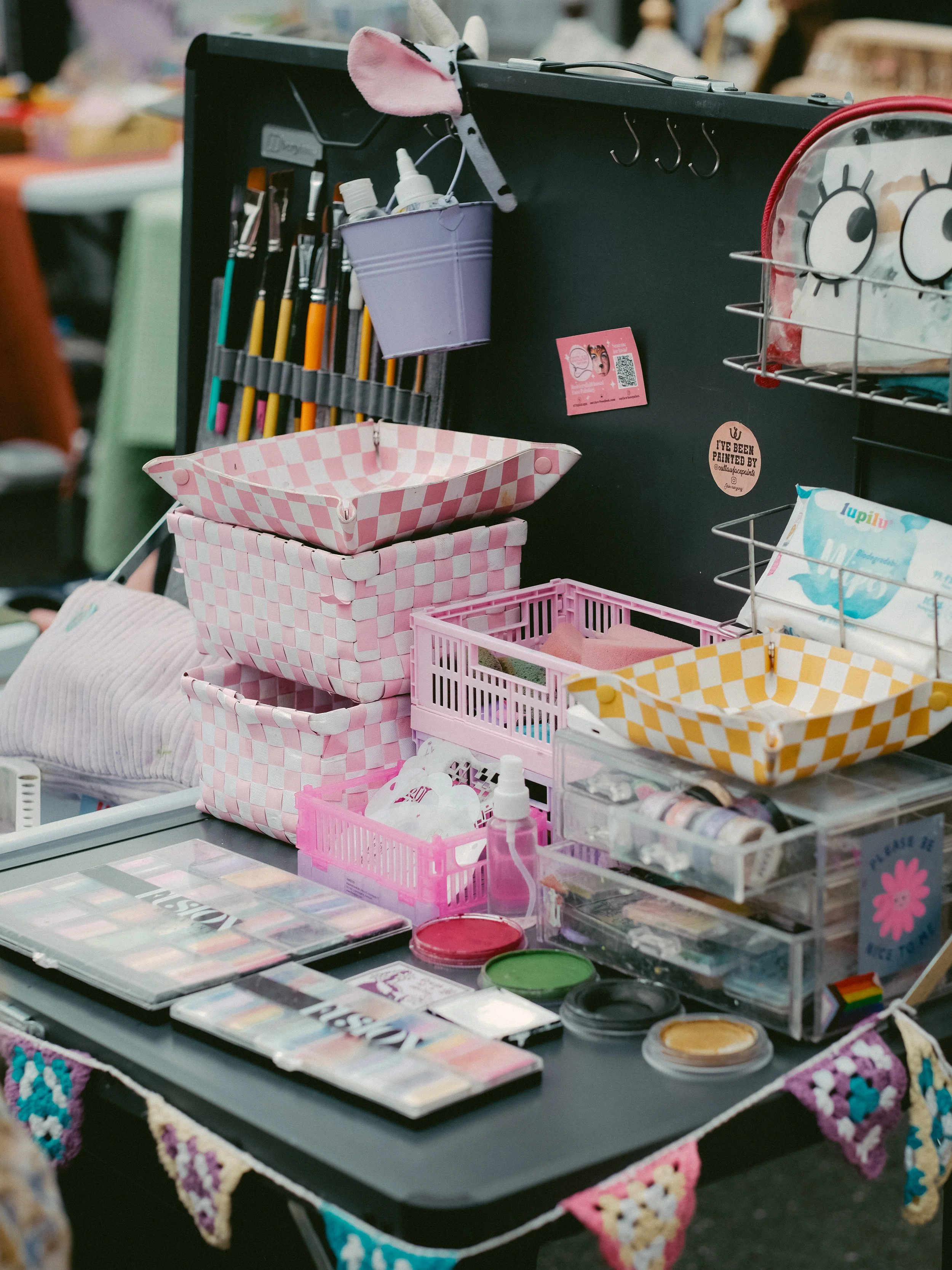 Craft supplies on a black table at a market stall, including colorful paints, patterned baskets, spray bottles, and decorative items.