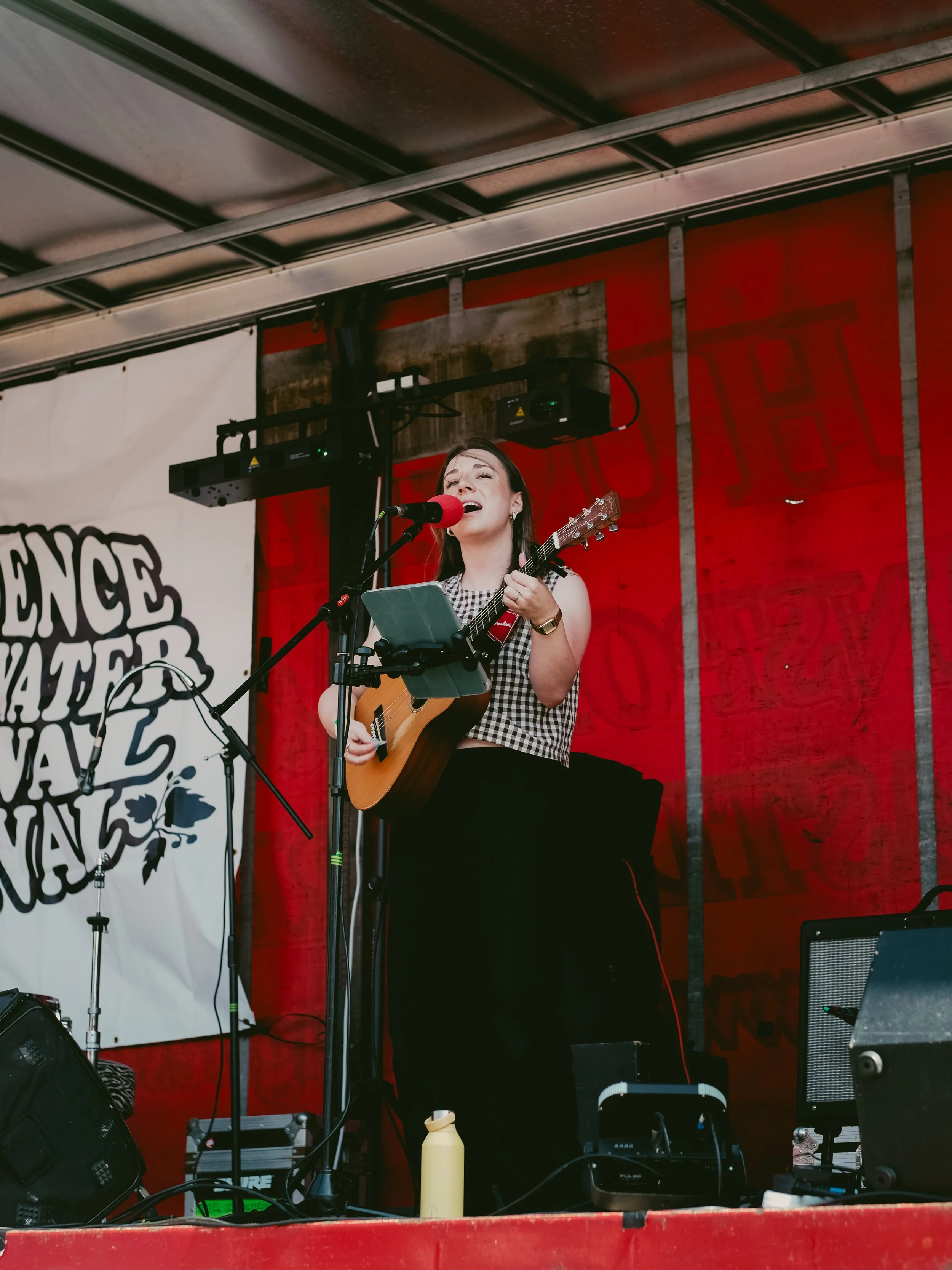 A woman singing and playing guitar on stage with a red backdrop, microphone, and speakers at a music festival.